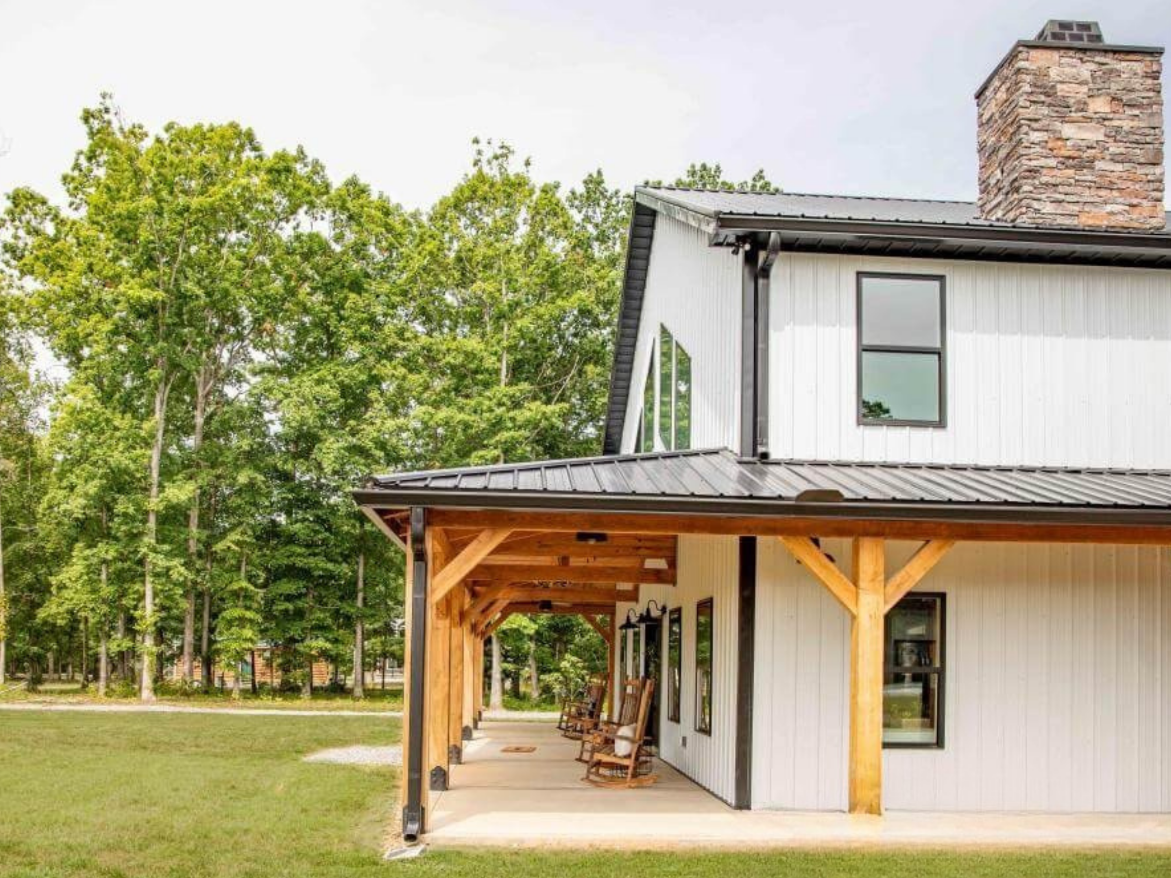 Side view of a modern white house with black window frames, a stone chimney, and a wooden porch with rocking chairs, surrounded by green grass and trees in the background.
