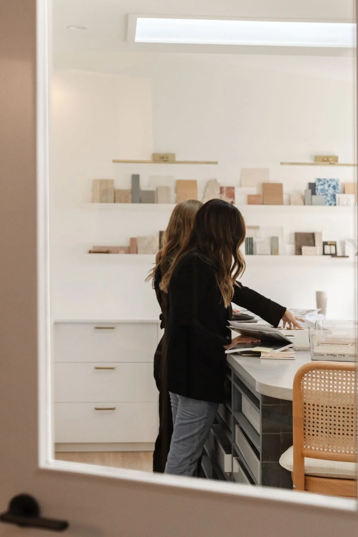 Two women with long hair, one with blonde and one with brunette, are looking through magazines or catalogs at a kitchen island in a modern, minimalist kitchen.