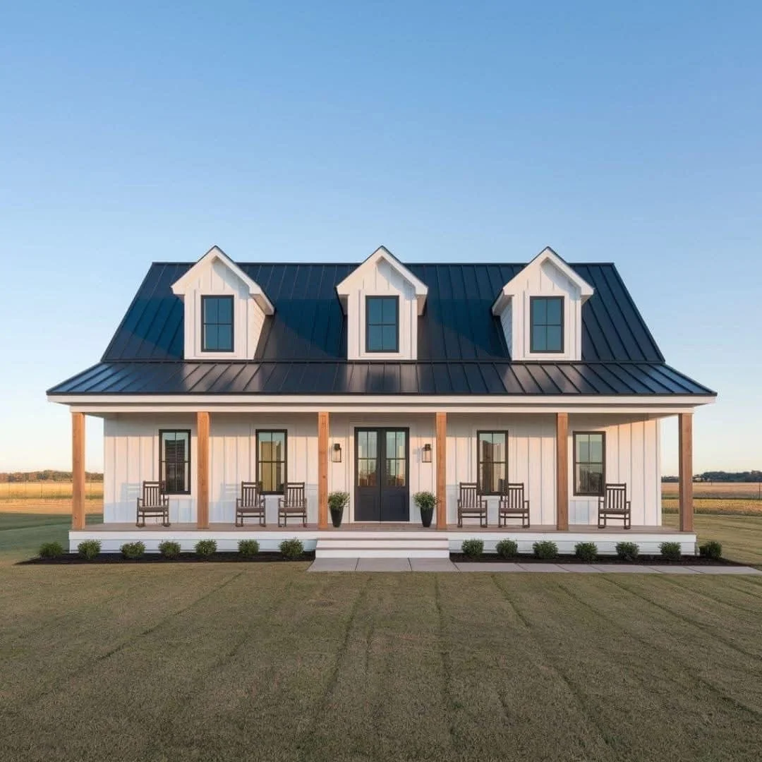 A modern farmhouse with white vertical siding, a black metal roof, three dormer windows, covered front porch with six chairs and two potted plants, set in a large grassy yard under a clear blue sky.