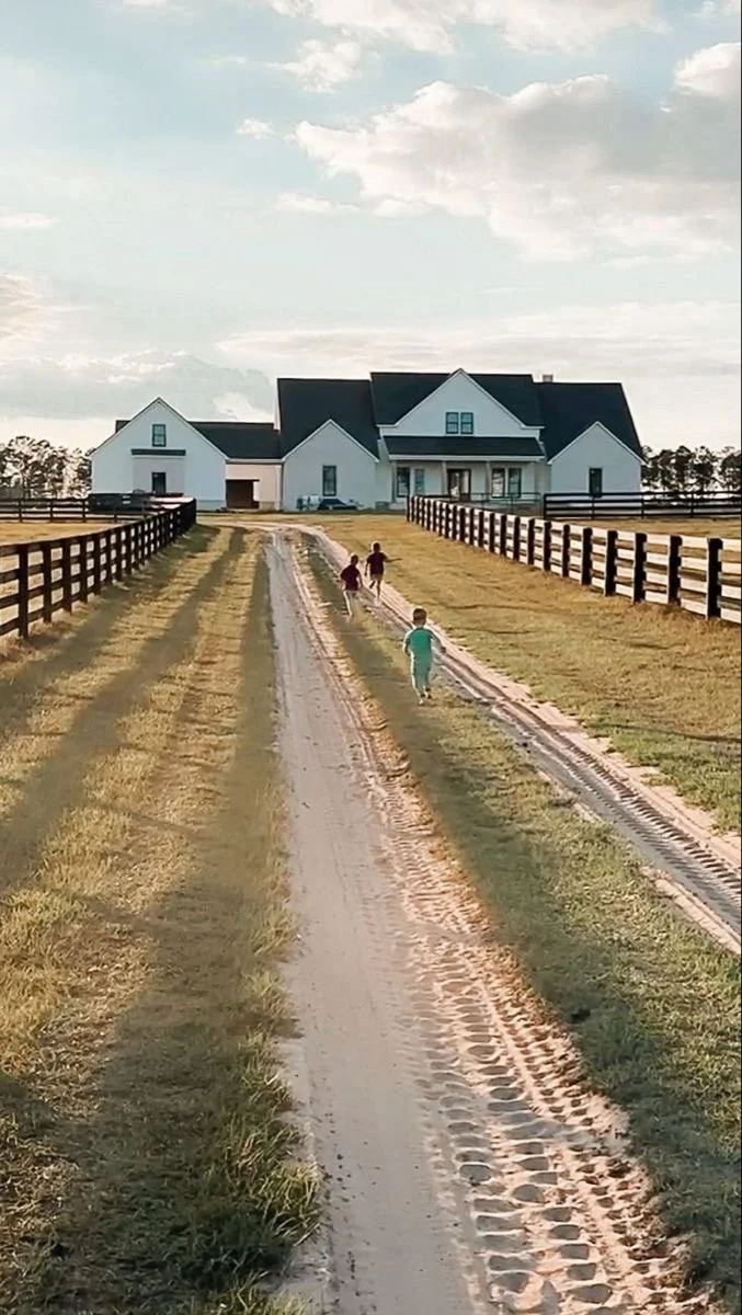 Three children walking along a dirt path toward a large white house with black roof, fenced sides, grassy surroundings, under a partly cloudy sky, in a rural setting.
