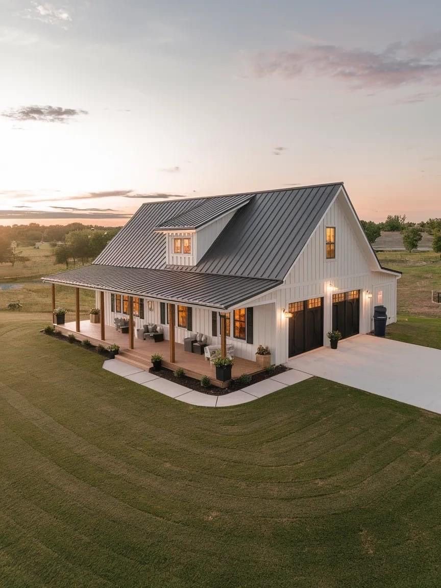 A modern farmhouse with a metal roof, white exterior, covered porch with seating, and a two-car garage, situated on a spacious lawn at sunset.