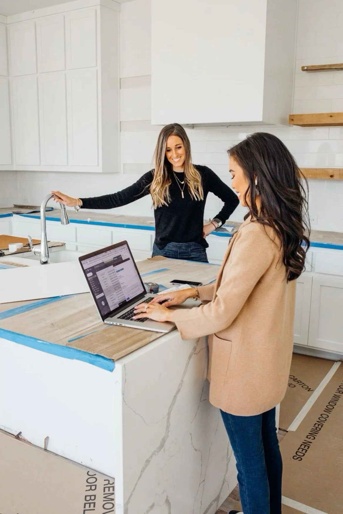 Two women in a kitchen under construction; one using a laptop, the other standing and smiling.