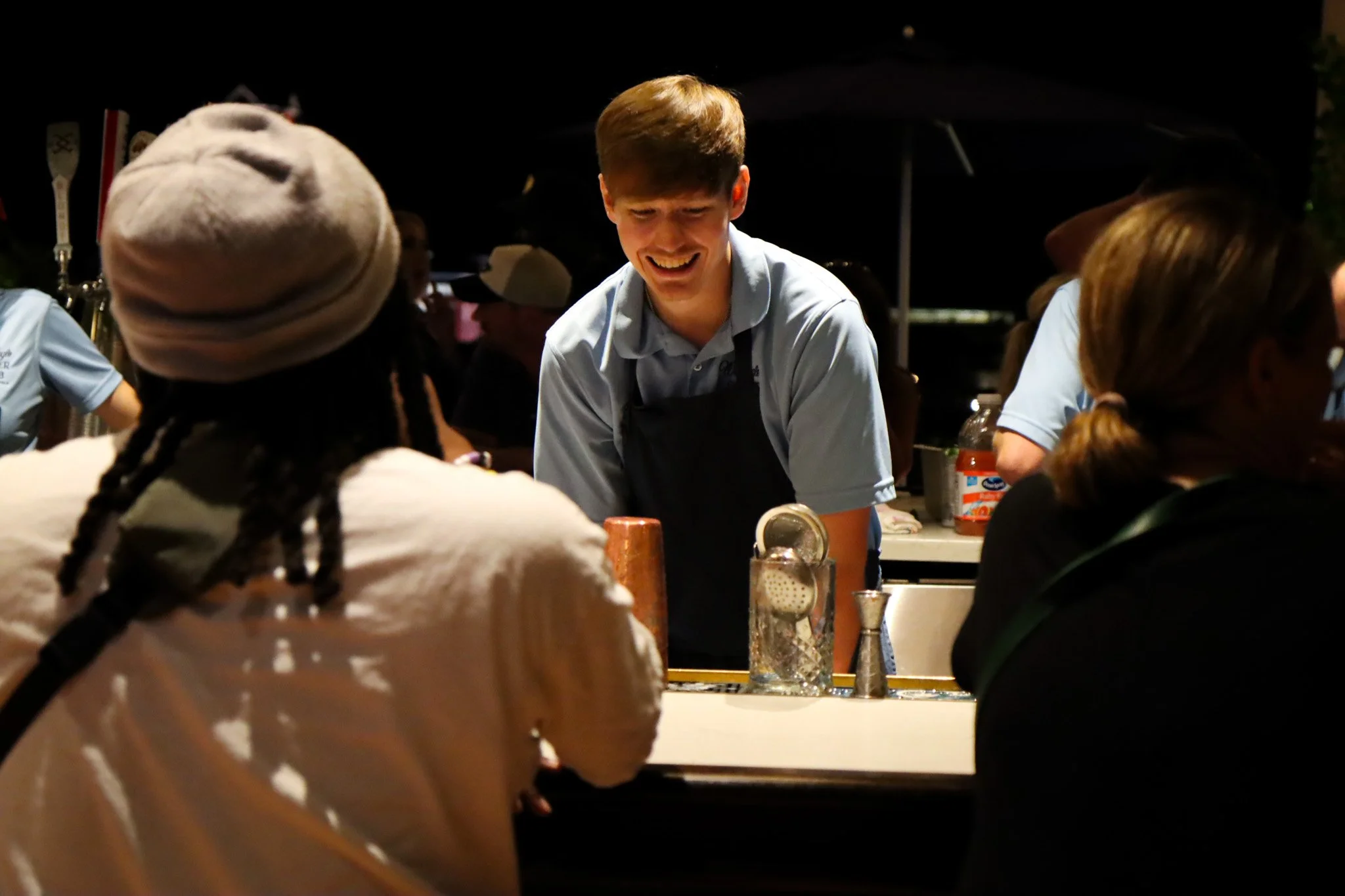 A young man serving at an outdoor restaurant during nighttime, smiling and engaging with customers.
