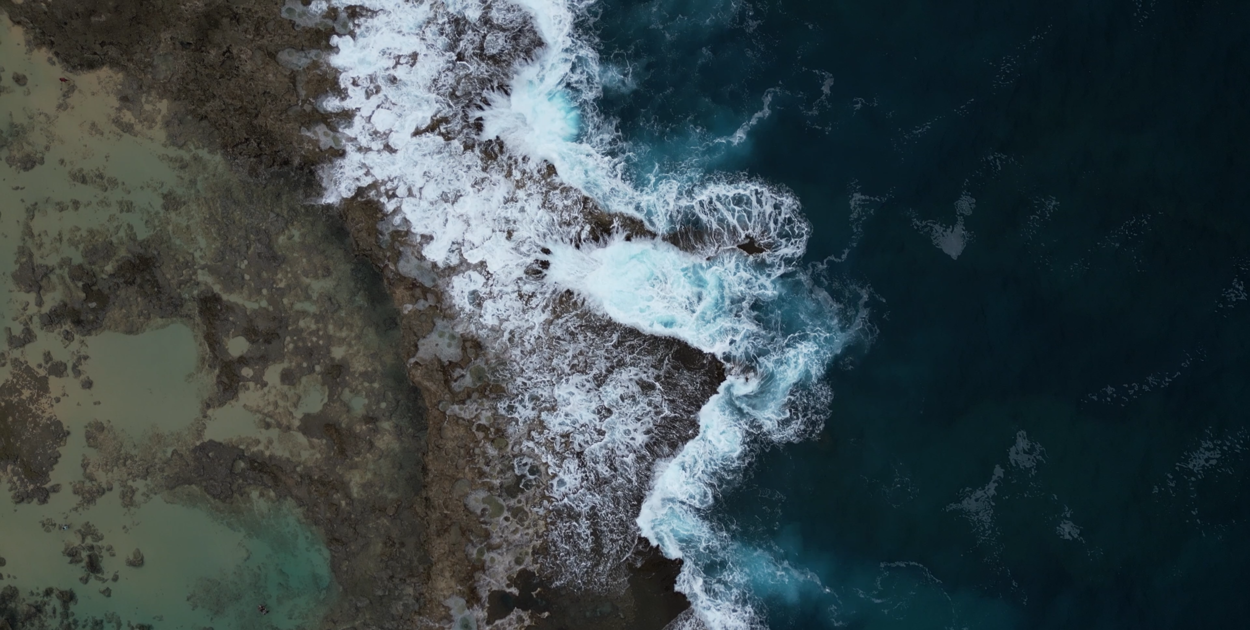 Aerial view of ocean waves crashing against a rocky shoreline with dark blue water.