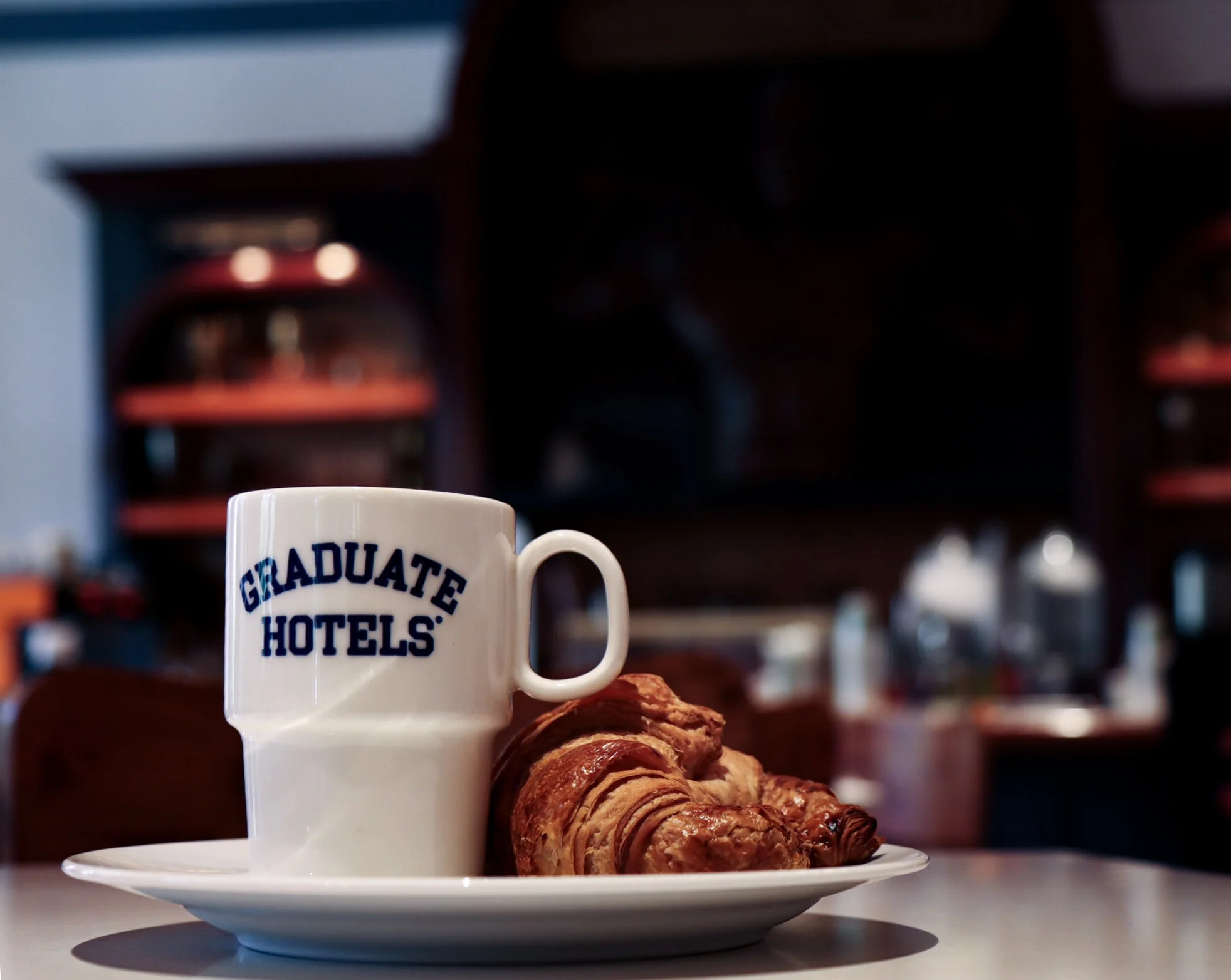A white mug with blue text that reads 'Graduate Hotels' on a plate with a croissant, set on a table. The background is out of focus.