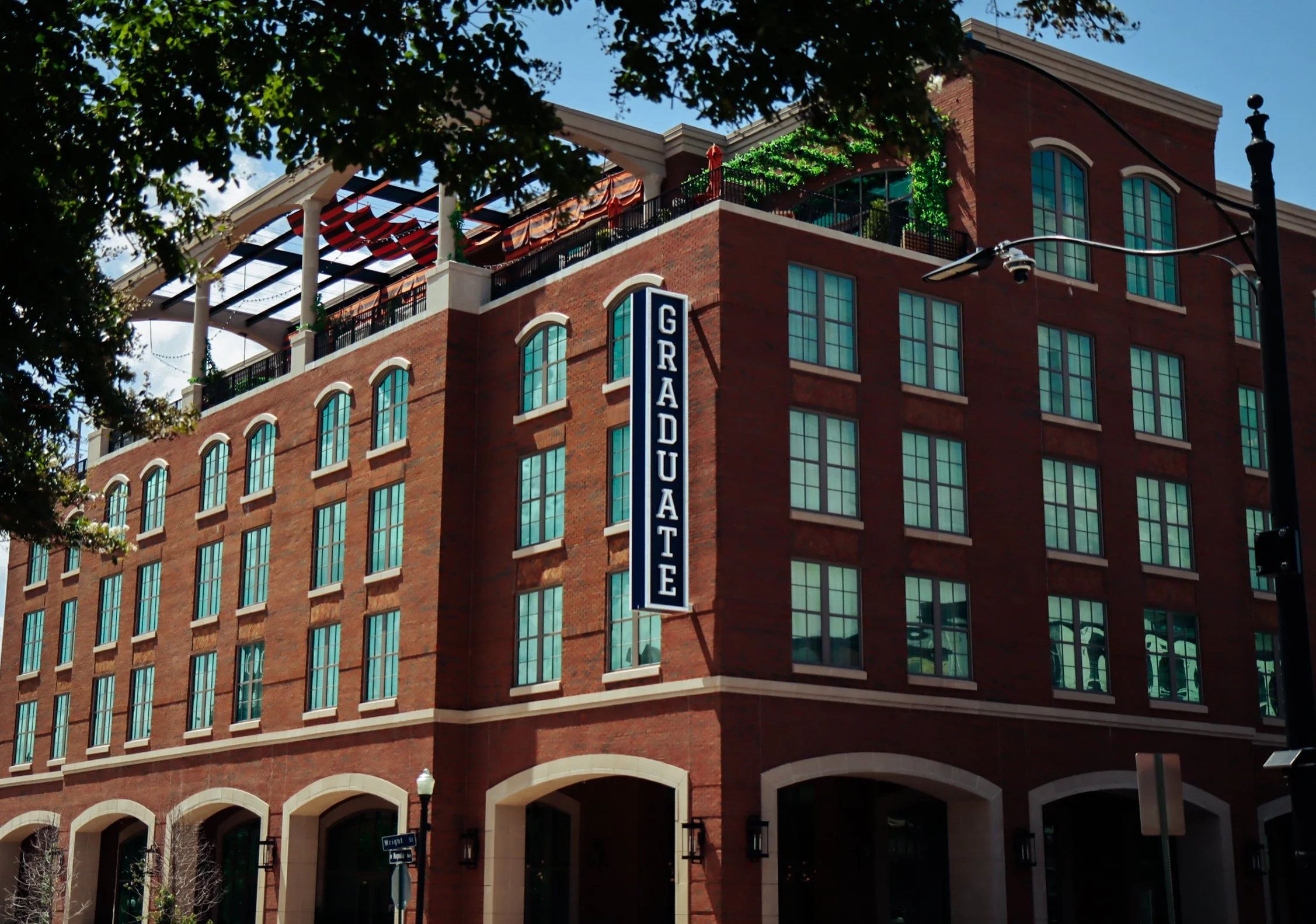 A brick building with multiple large windows and a vertical sign that reads 'GRADUATE'. The building has a rooftop area with greenery and outdoor furniture, partially shaded by a tree.