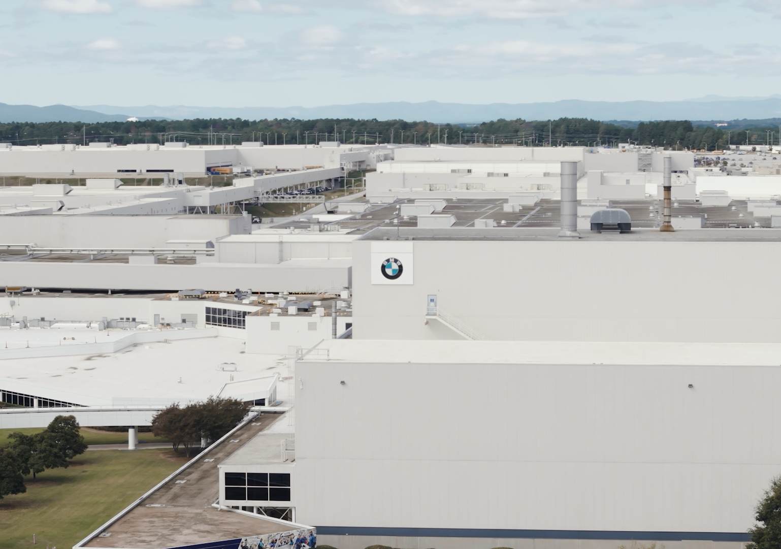 Large manufacturing or industrial facility with white buildings, a BMW logo, and smoke stacks, situated in a semi-rural area with trees and mountains in the background.
