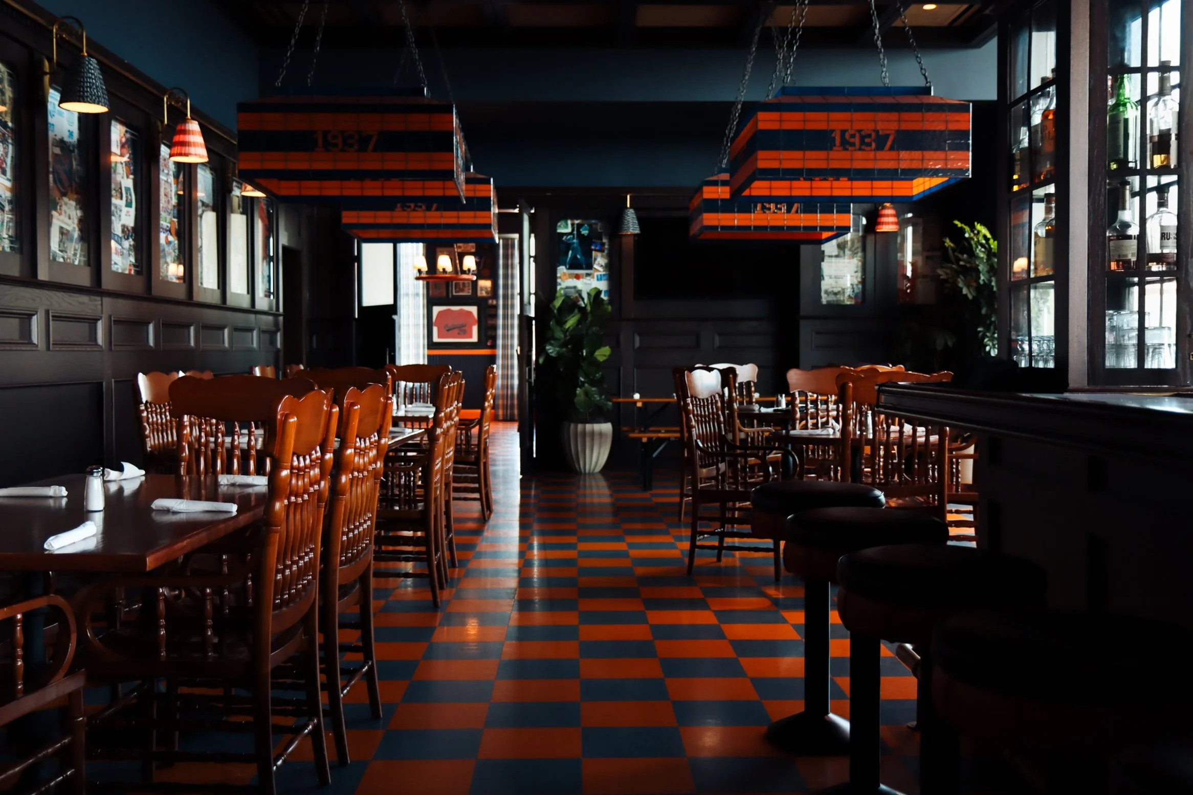 Interior of a restaurant with dark wood furniture, checkered orange and black floors, hanging light fixtures, and windows on the right side.