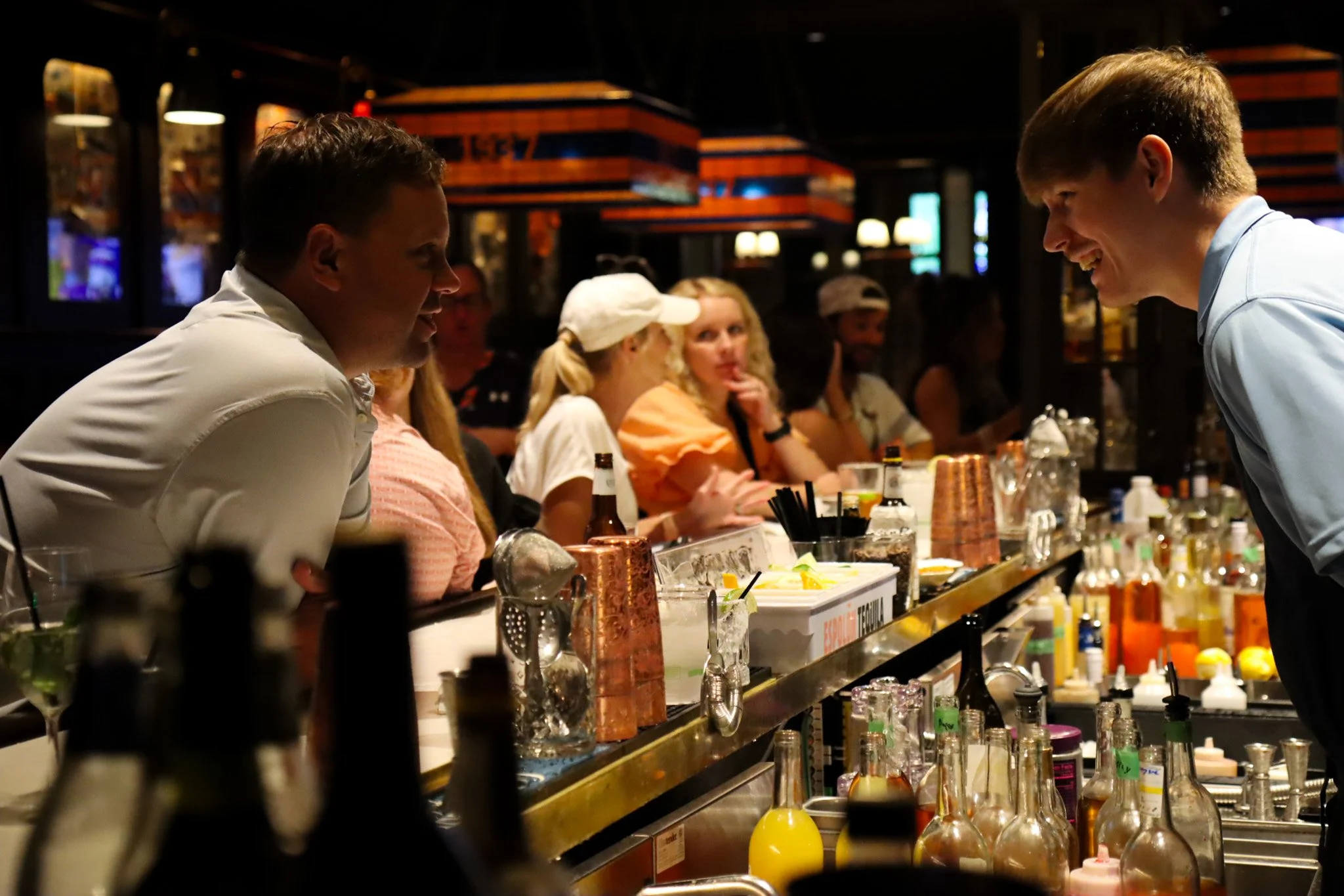 Two bartenders facing each other, smiling in a busy bar with patrons in the background.