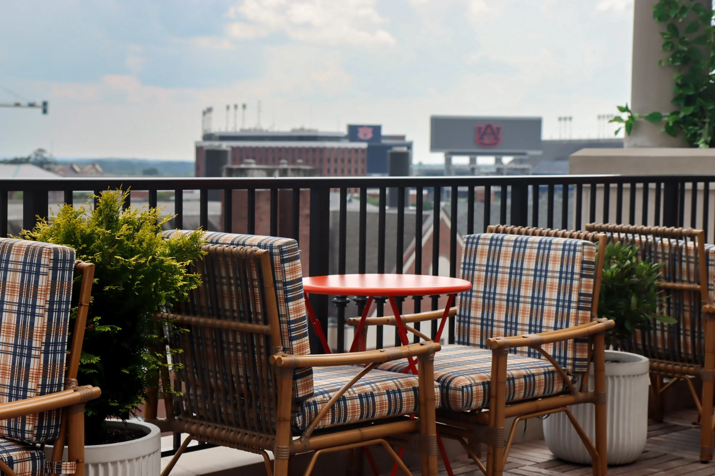 Outdoor balcony patio with plaid cushioned chairs, a small red table, potted plants, and city buildings in the background
