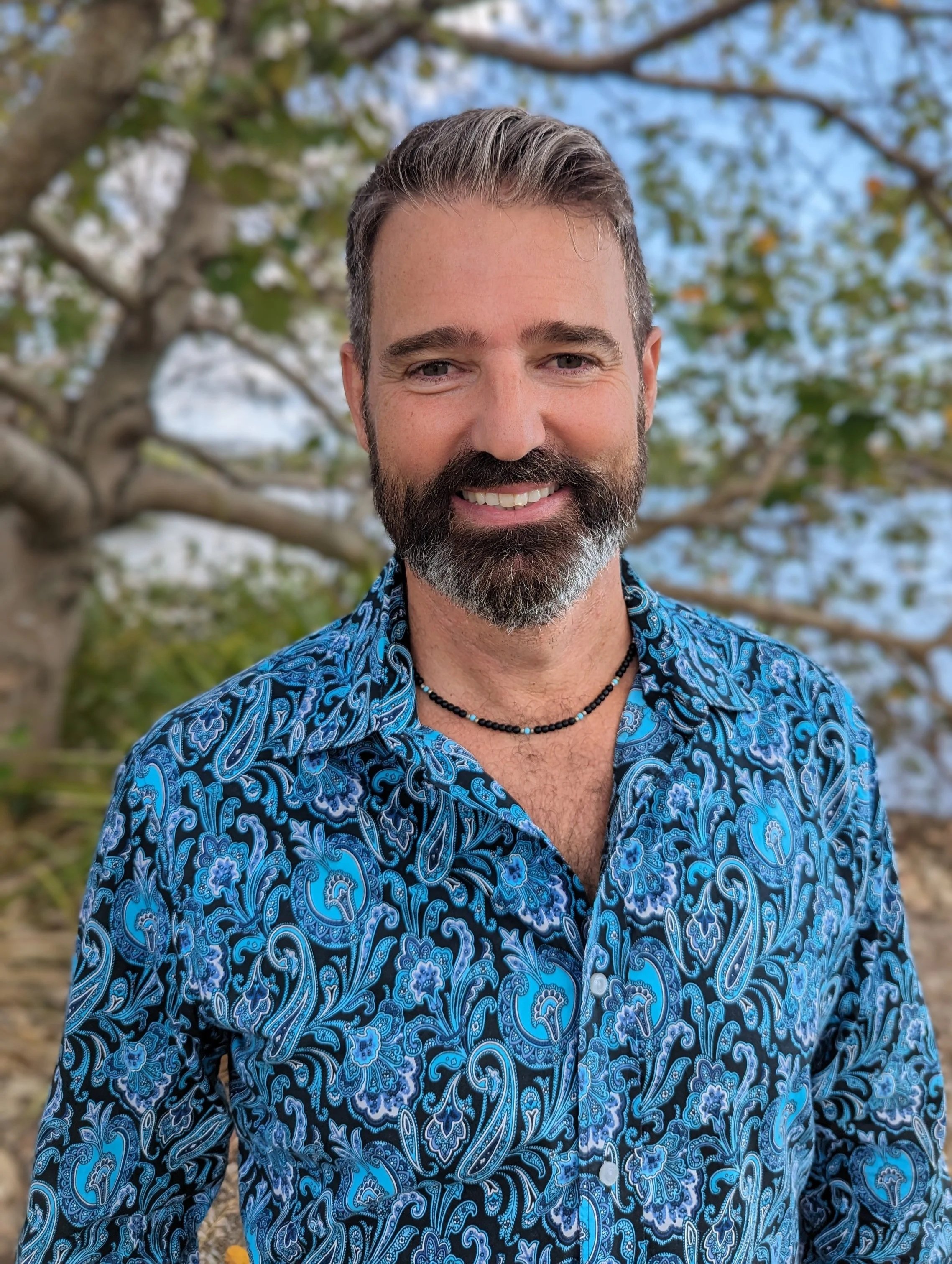 Anthony Spark is a smiling celebrant in Mooloolaba wearing a blue patterned shirt and a black beaded necklace outdoors near a tree with a blue sky in the background.