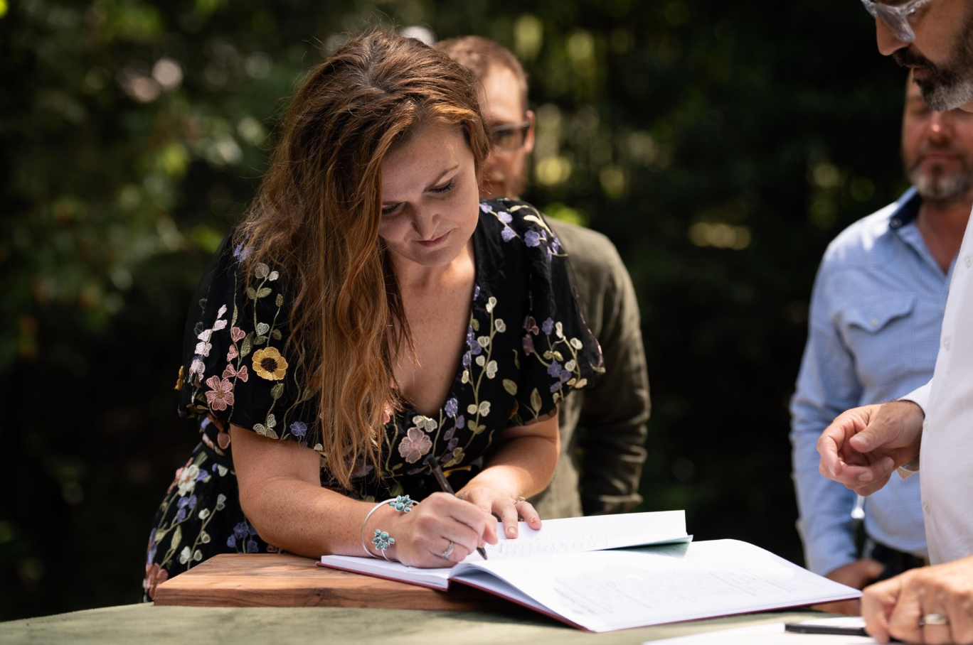 A bride with long, wavy red hair signs a wedding certificate outdoors while three men watch, her new husband, father-in-law and celebrant- Anthony Spark