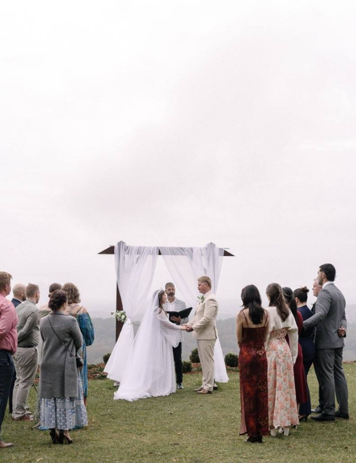 Wedding ceremony outdoors with bride, groom, and guests under a white draped canopy.