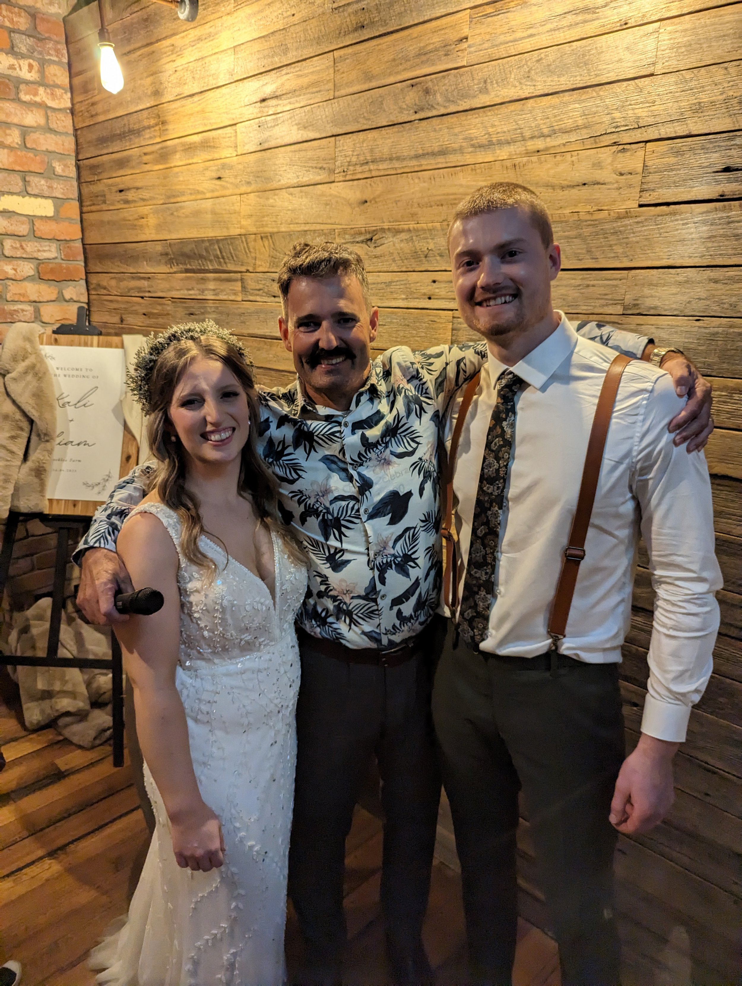 A wedding reception with bride, groom and celebrant- Anthony Spark. They are smiling and standing close together against a wooden wall with a brick section and a light bulb hanging from the ceiling.