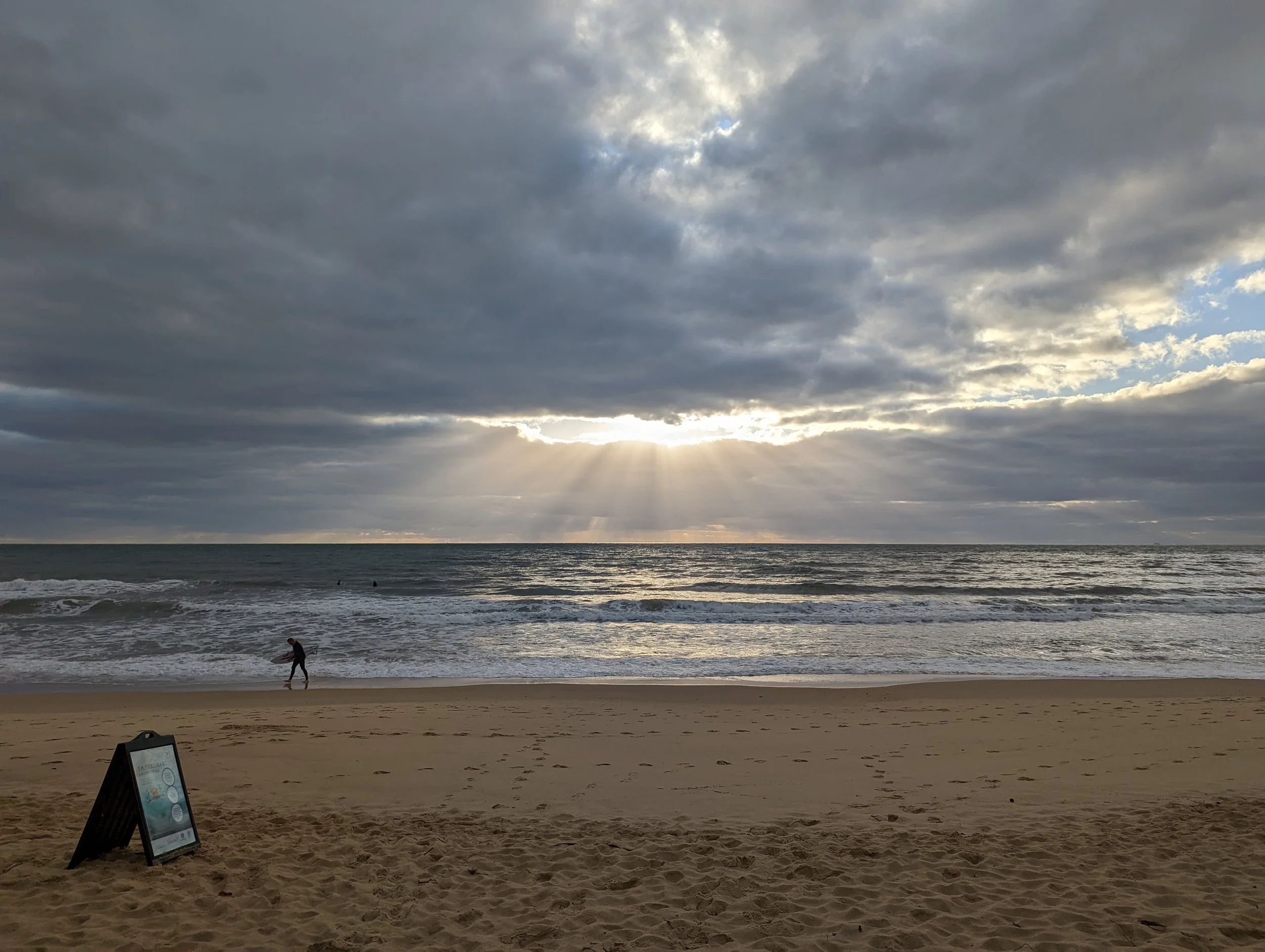A beach scene with cloudy sky, sun rays peeking through the clouds, a sandy shore, and the ocean with small waves. A person is walking along the shoreline near a signboard on the sand.