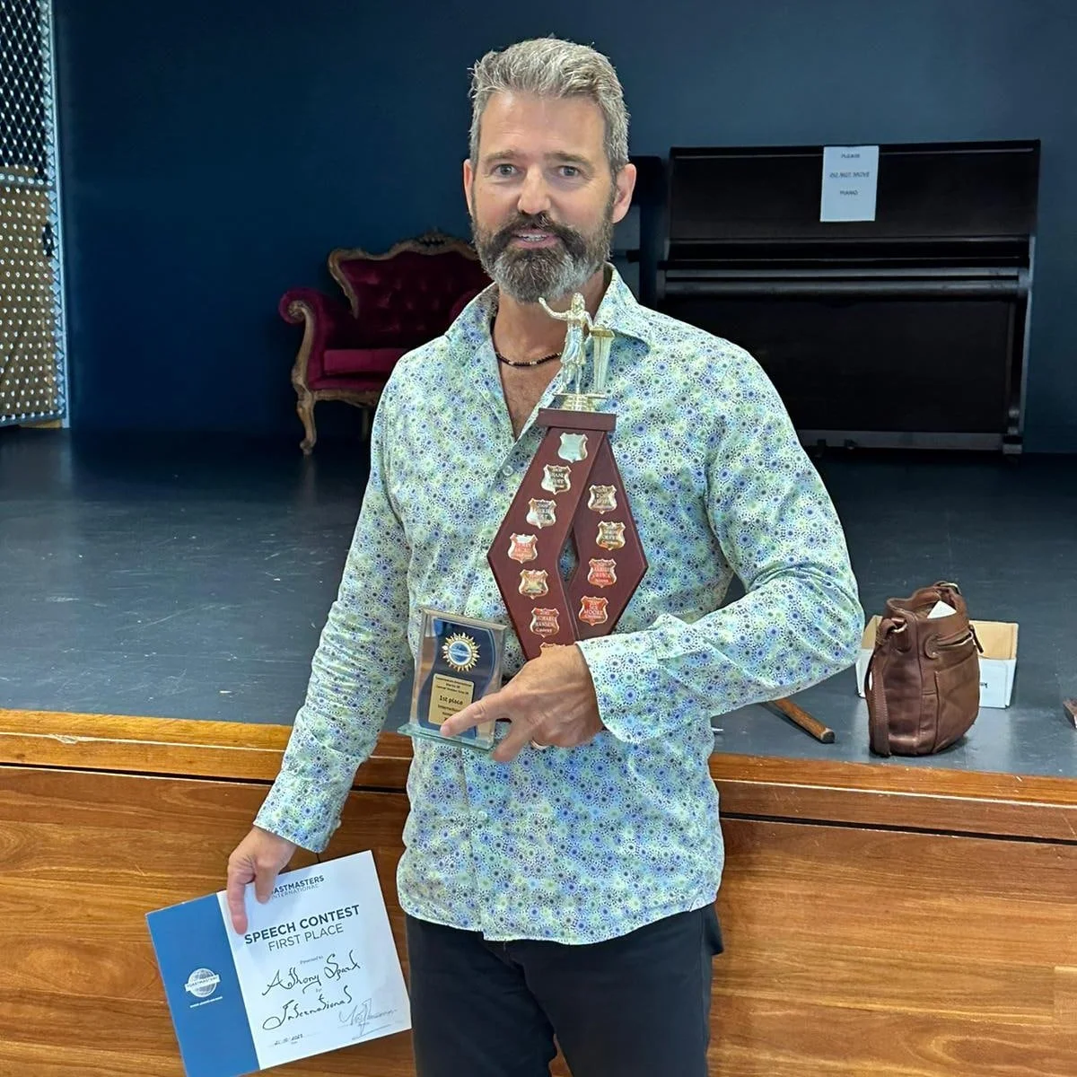 Anthony Spark after winning a Toastmasters contest holding a trophy and a first-place speech contest certificate. He stands on a stage with a dark background and a vintage red and gold sofa. Items like a brown bag and a box are on the stage.