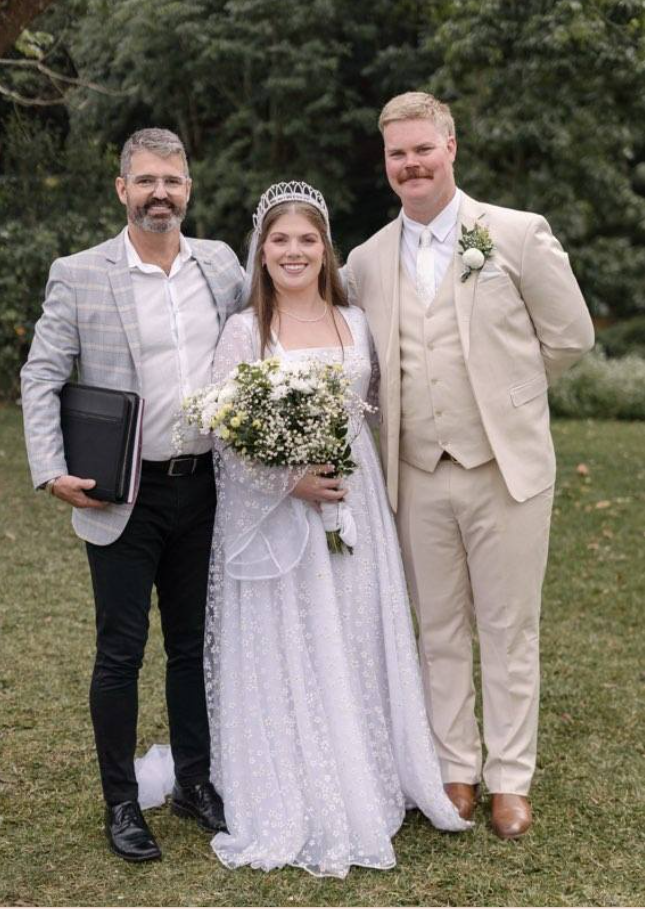 Three people standing outdoors on grass; a bride in a white wedding dress with a bouquet, a groom in a cream suit, and an officiant with a folder, all smiling.