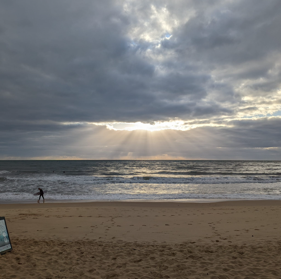 Sunlight breaking through dark storm clouds over the ocean, with a sandy beach in the foreground and a person carrying a surfboard walking along the shoreline.