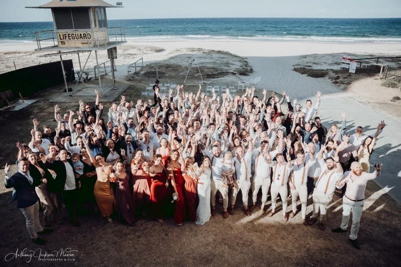 Group of people celebrating a wedding outdoors near a sandy beach with a lifeguard tower in the background.