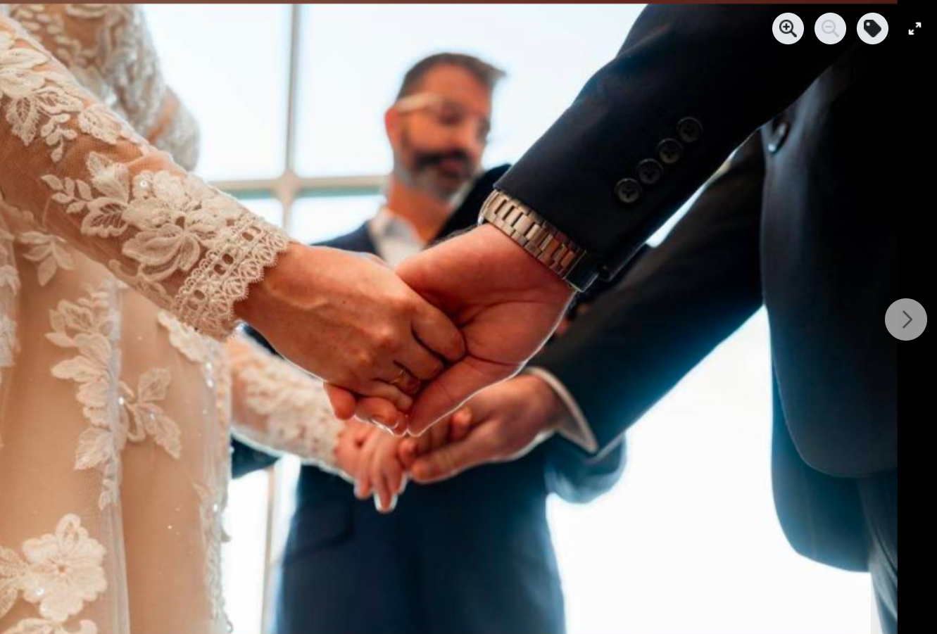 A close-up of a wedding ceremony where a bride and groom hold hands during their vows, with officiant, Anthony Spark in the background.