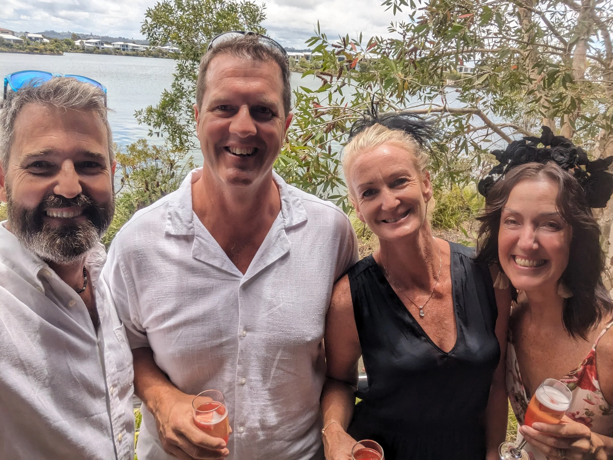 Four people smiling at a gathering near a body of water, holding glasses with drinks, with trees and water in the background.
