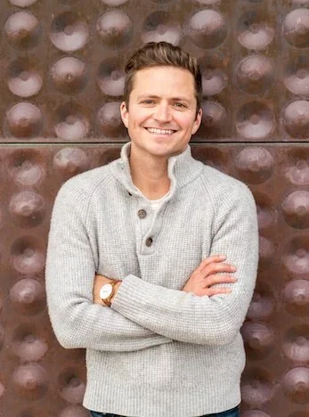 A young man with light brown hair, smiling, wearing a light gray sweater, standing in front of a textured metallic background with circular patterns.
