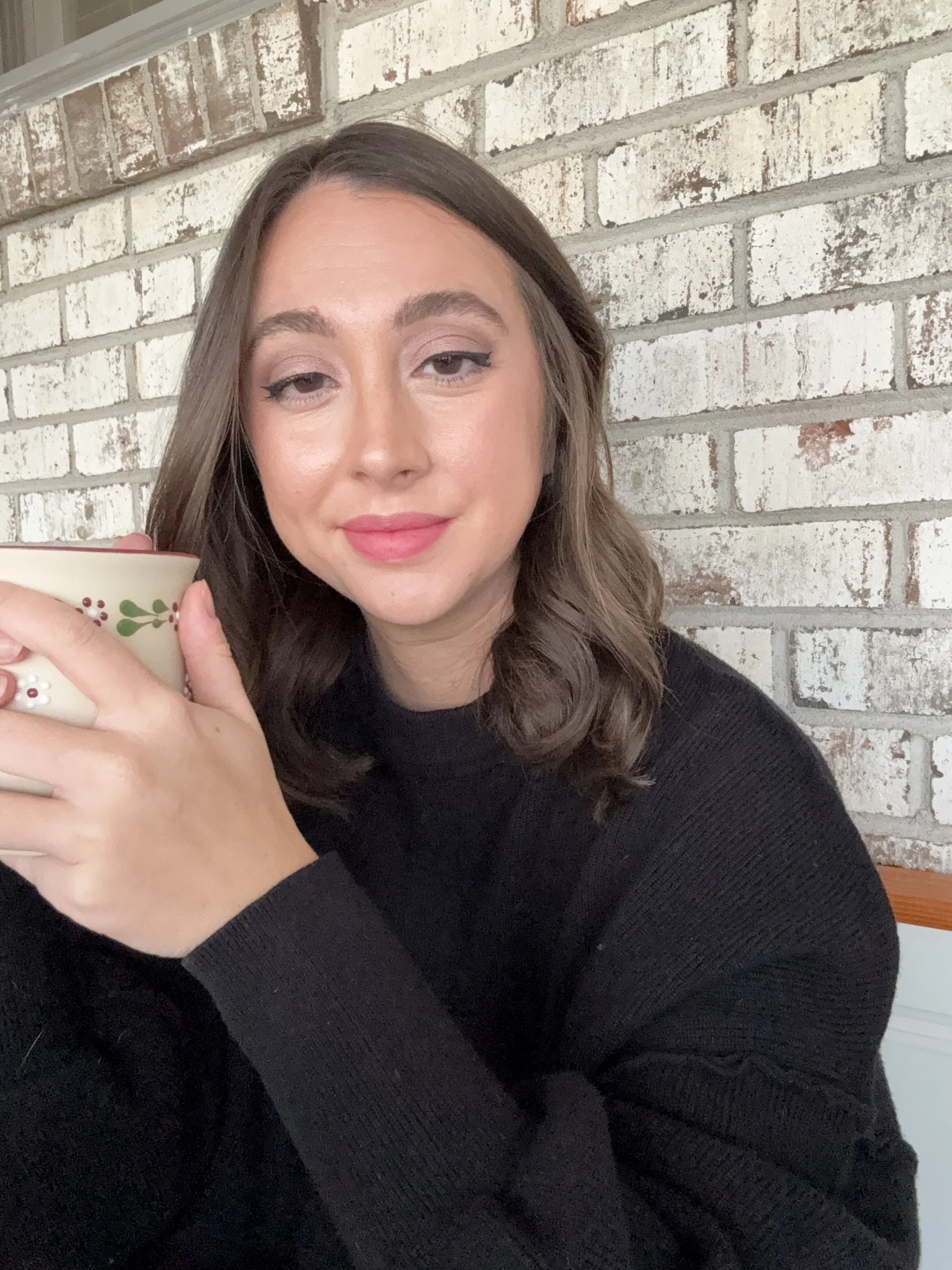 A woman with shoulder-length brown hair and light makeup, wearing a black sweater, holding a decorated mug, sitting in front of a white brick wall.