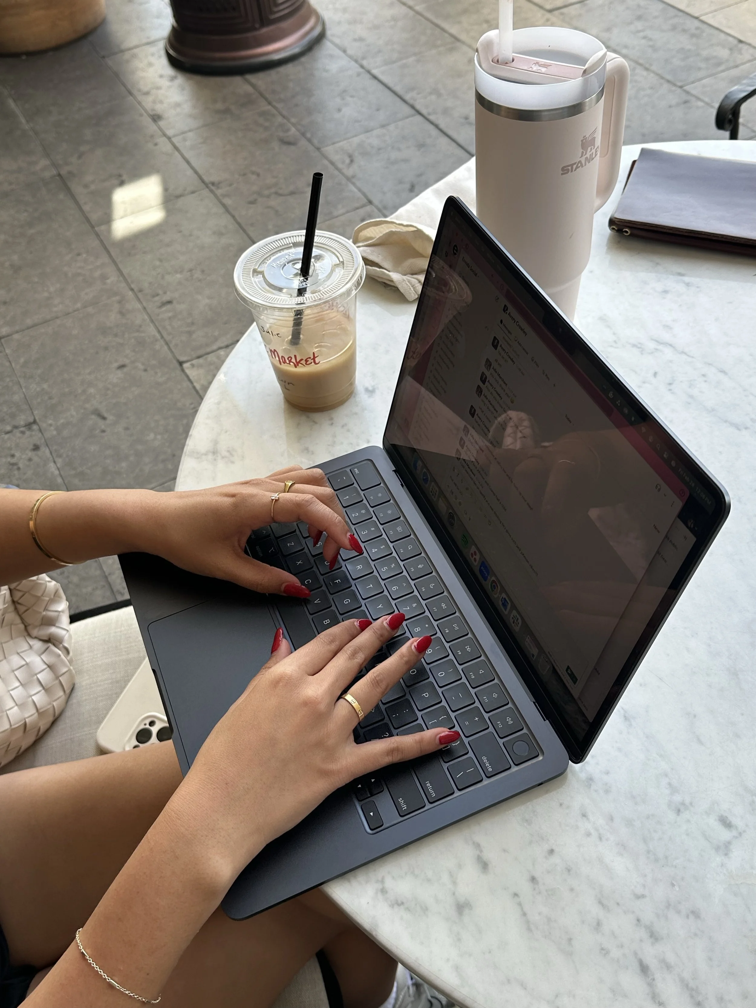 Person working on a laptop at a table outside, with iced coffee and a thermal mug also on the table.