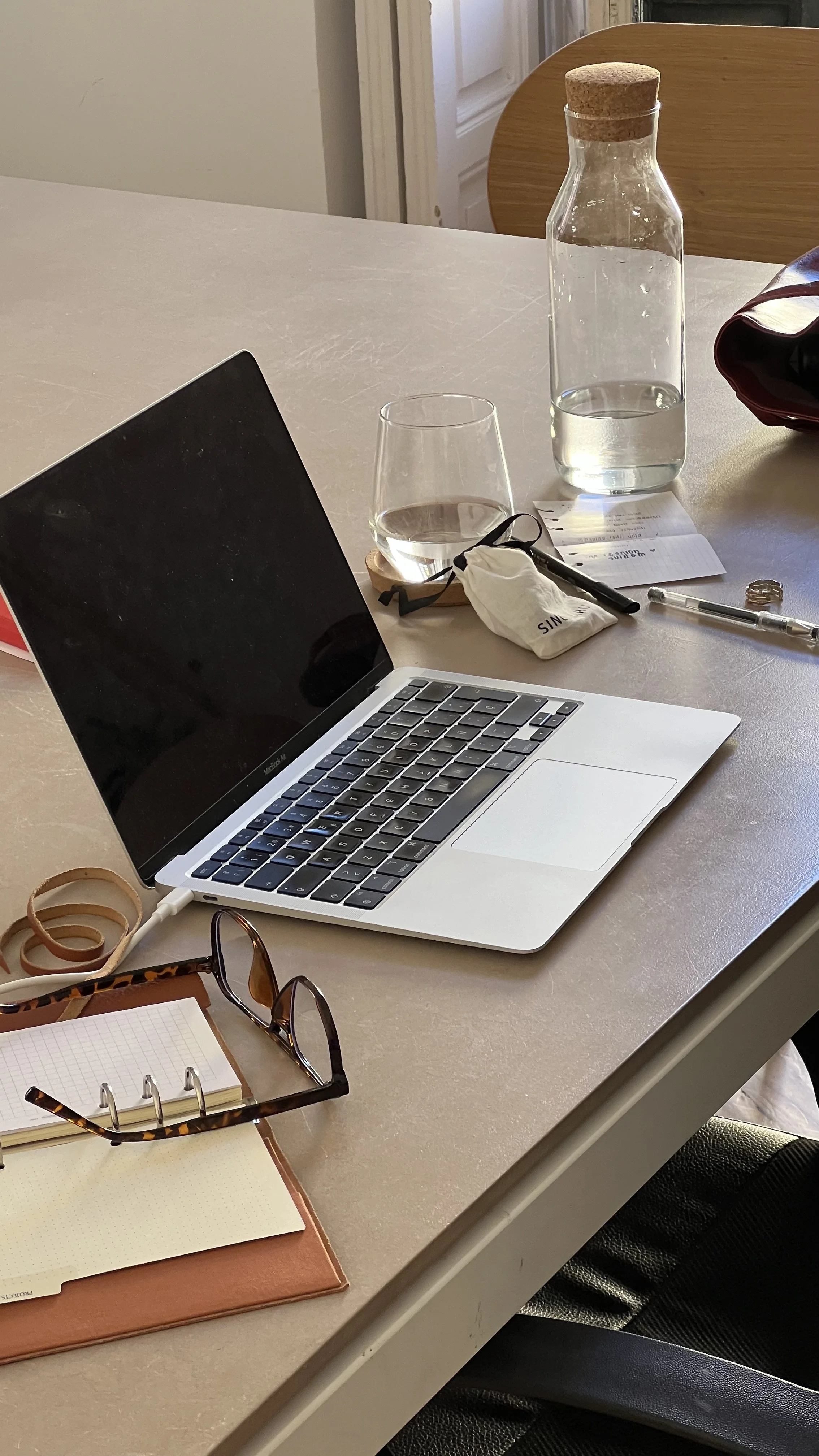 A cluttered desk with a MacBook, eyeglasses, a notebook, a wallet, a pen, a glass of water, a glass bottle of water, and a small pouch.