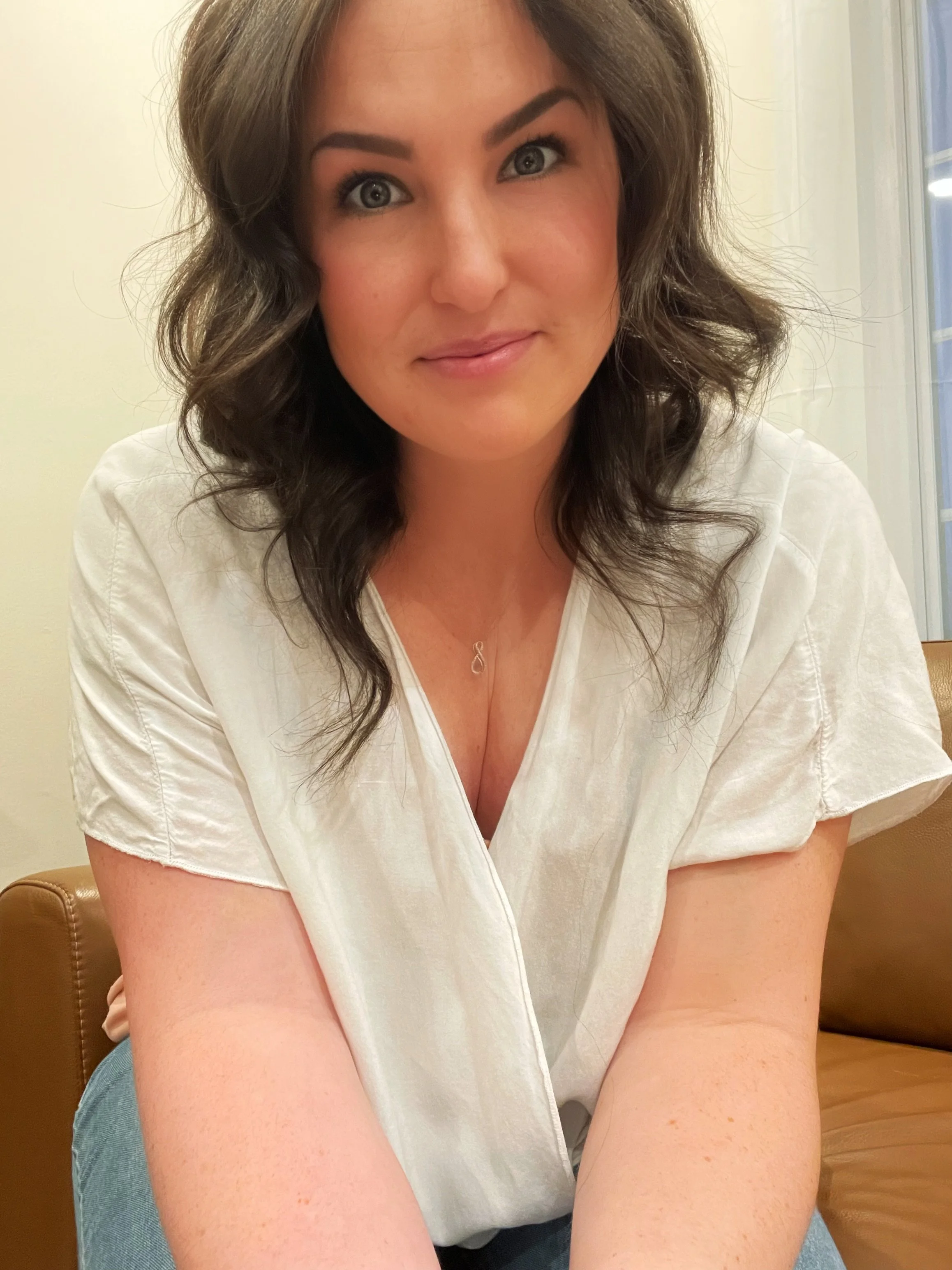 A woman with curly brown hair and blue eyes, wearing a white shirt and a delicate necklace, smiling softly, seated indoors near a window.