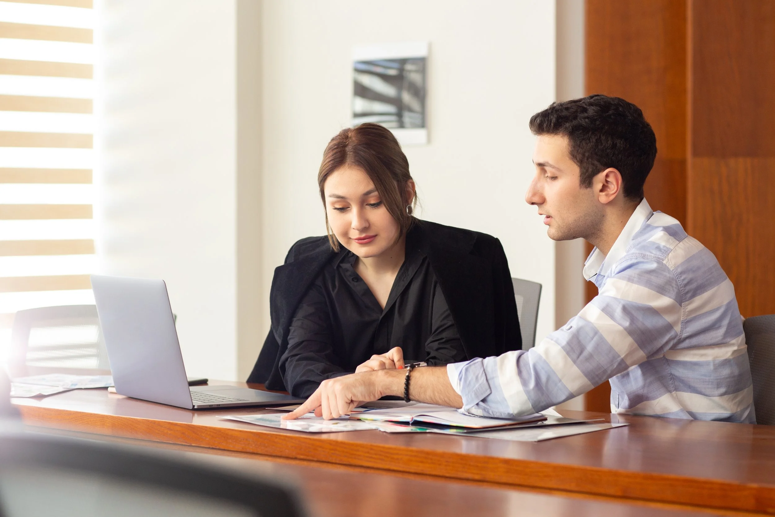 A man and woman sitting at a wooden table in an office, reviewing documents and looking at a laptop.