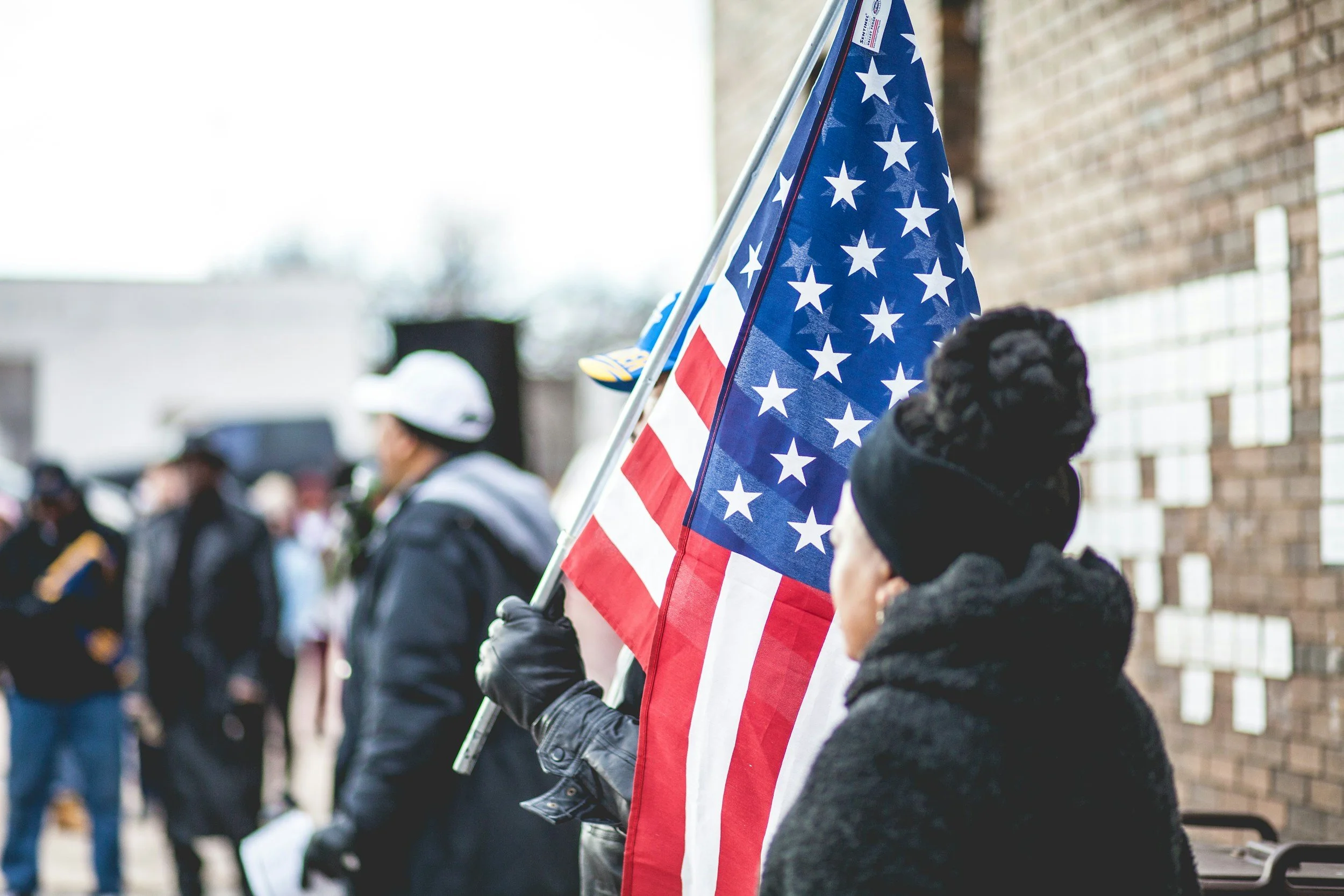Person holding an American flag during a public gathering or event near a brick wall.