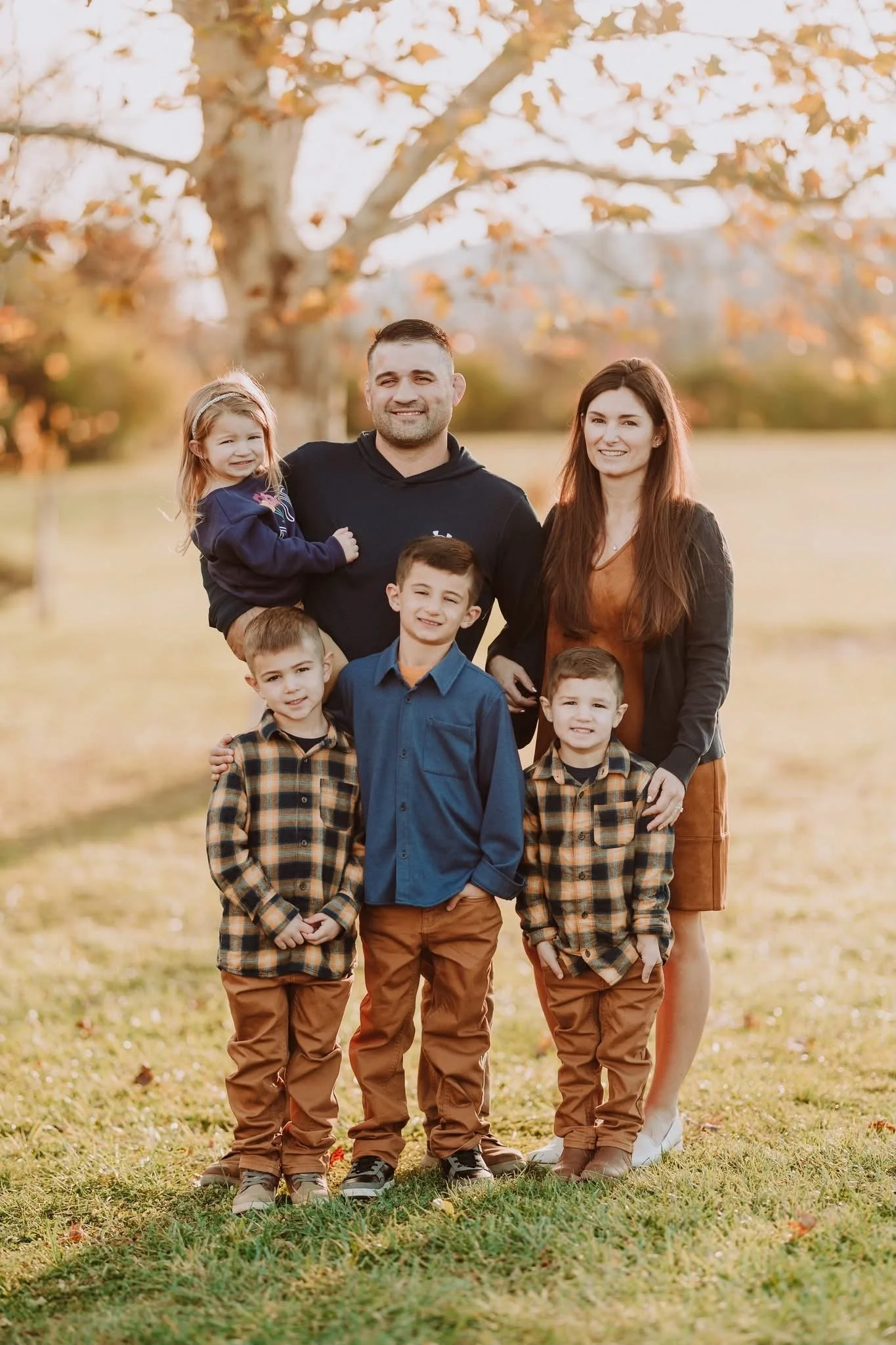 A family of seven standing outdoors on a fall day with trees and autumn leaves in the background.