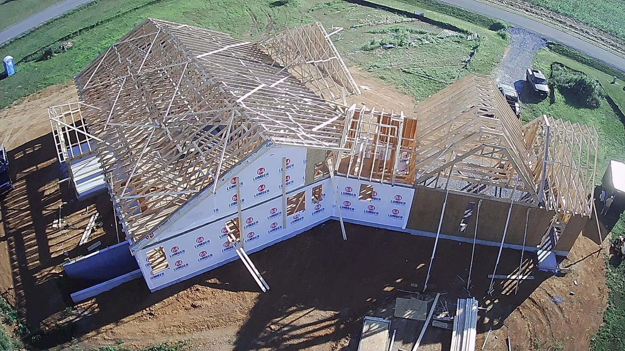 A house under construction with wooden framing on a dirt lot, partially covered with weather-resistant sheathing. The roof structure is in progress, with wooden trusses and beams forming the roof outline.
