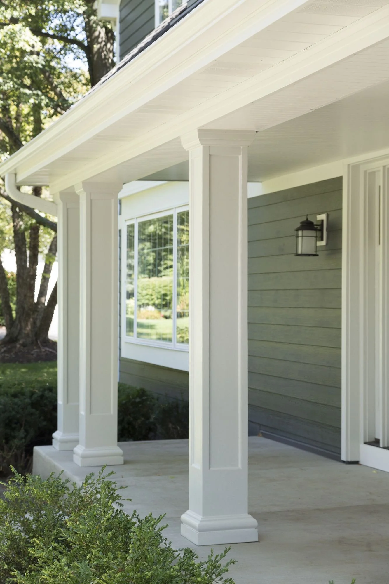 Close-up of the front porch of a house with white columns and a grey exterior wall, with a garden and a large tree in the background.