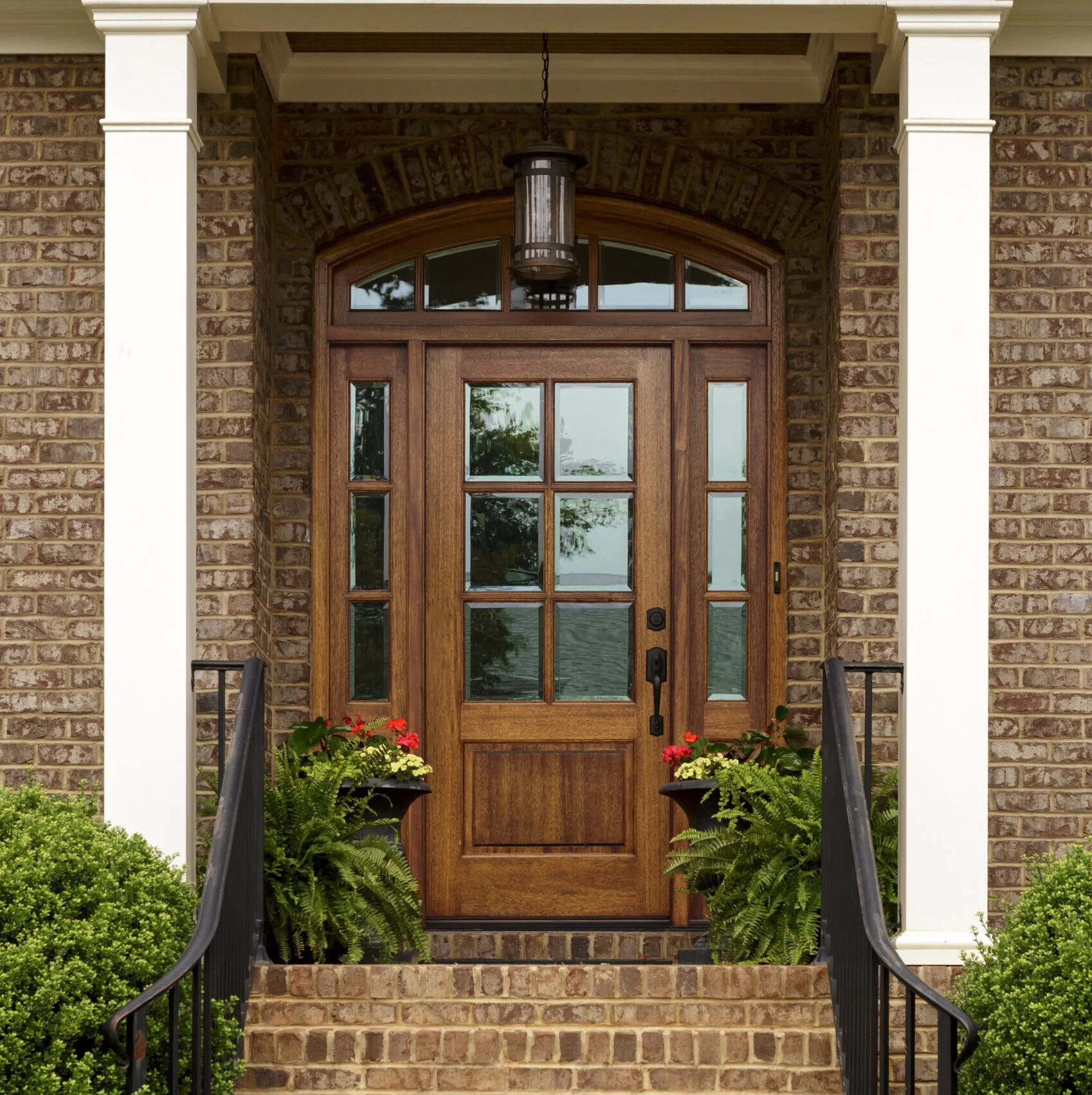 Wooden front door with glass panes, surrounded by brick walls and floral planters with greenery and red flowers, with black handrails on brick stairs.