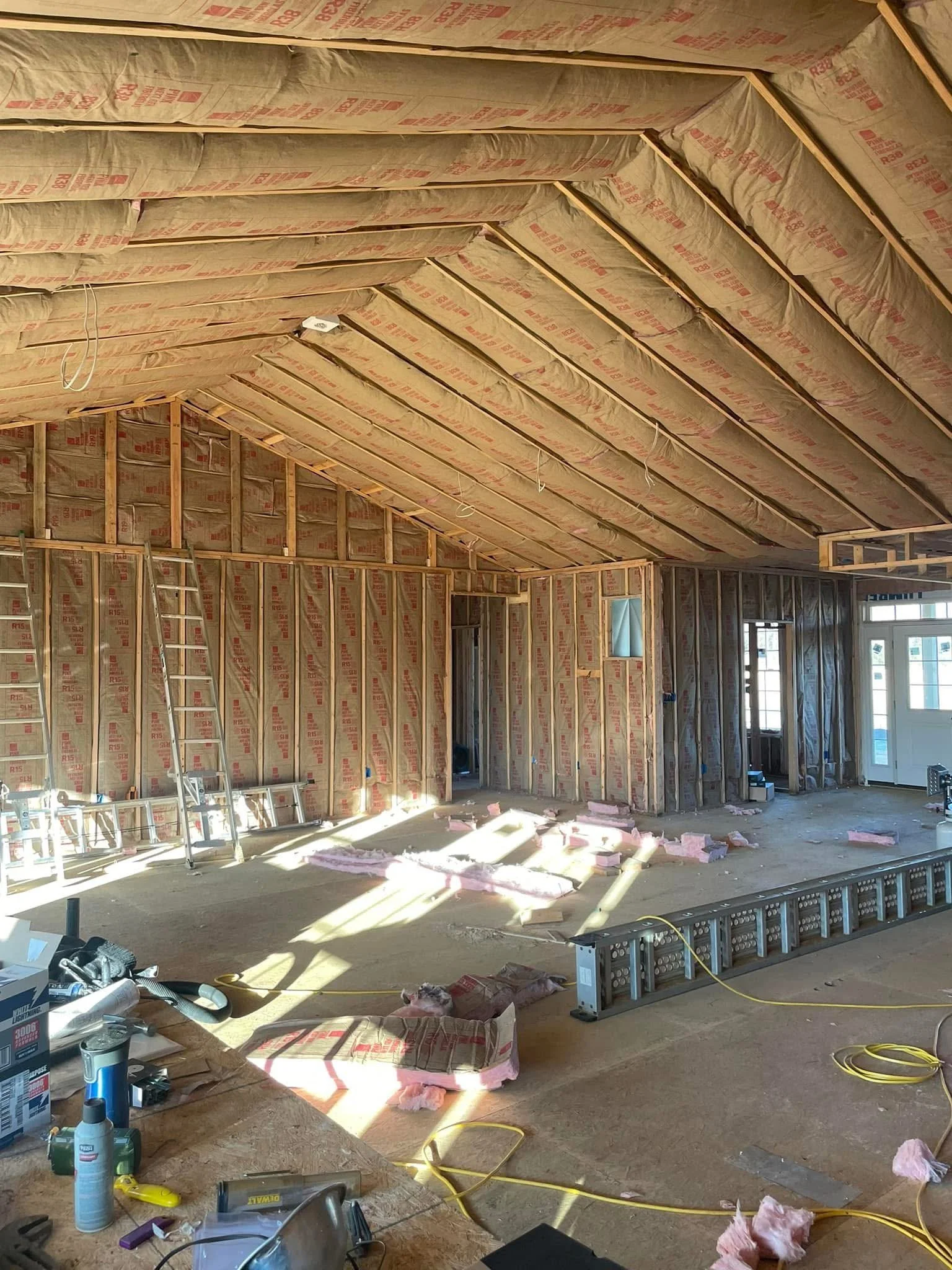 Interior of a house under construction with exposed wooden framing, insulation, and building materials scattered on the floor.