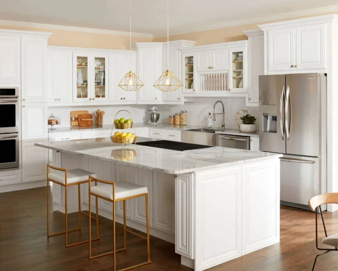 Bright white kitchen with marble island, stainless steel refrigerator, double ovens, and pendant lighting, featuring wooden stools with white cushions.