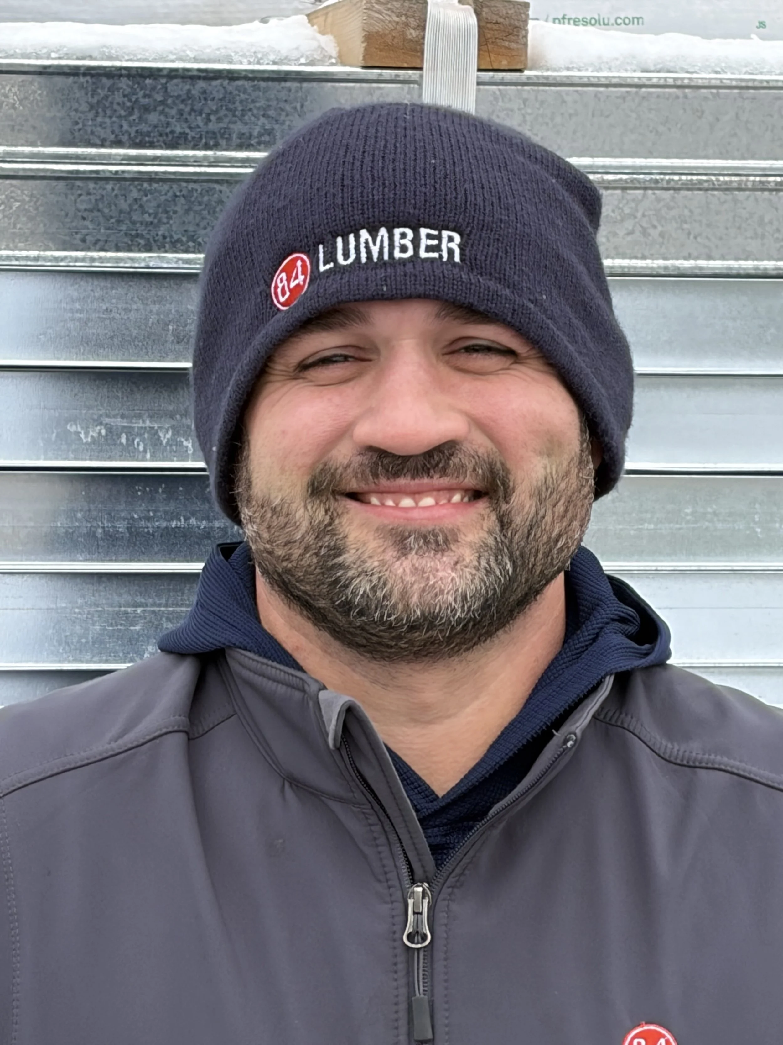 A man wearing a black beanie with '84 LUMBER' embroidered on it, smiling at the camera, standing in front of a metallic background.