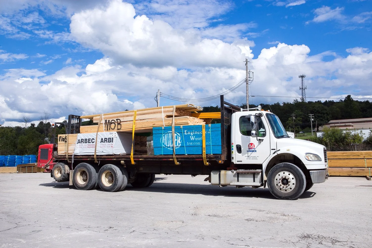 A white flatbed truck carrying lumber and pallets at a construction or hardware supply yard on a partly cloudy day.
