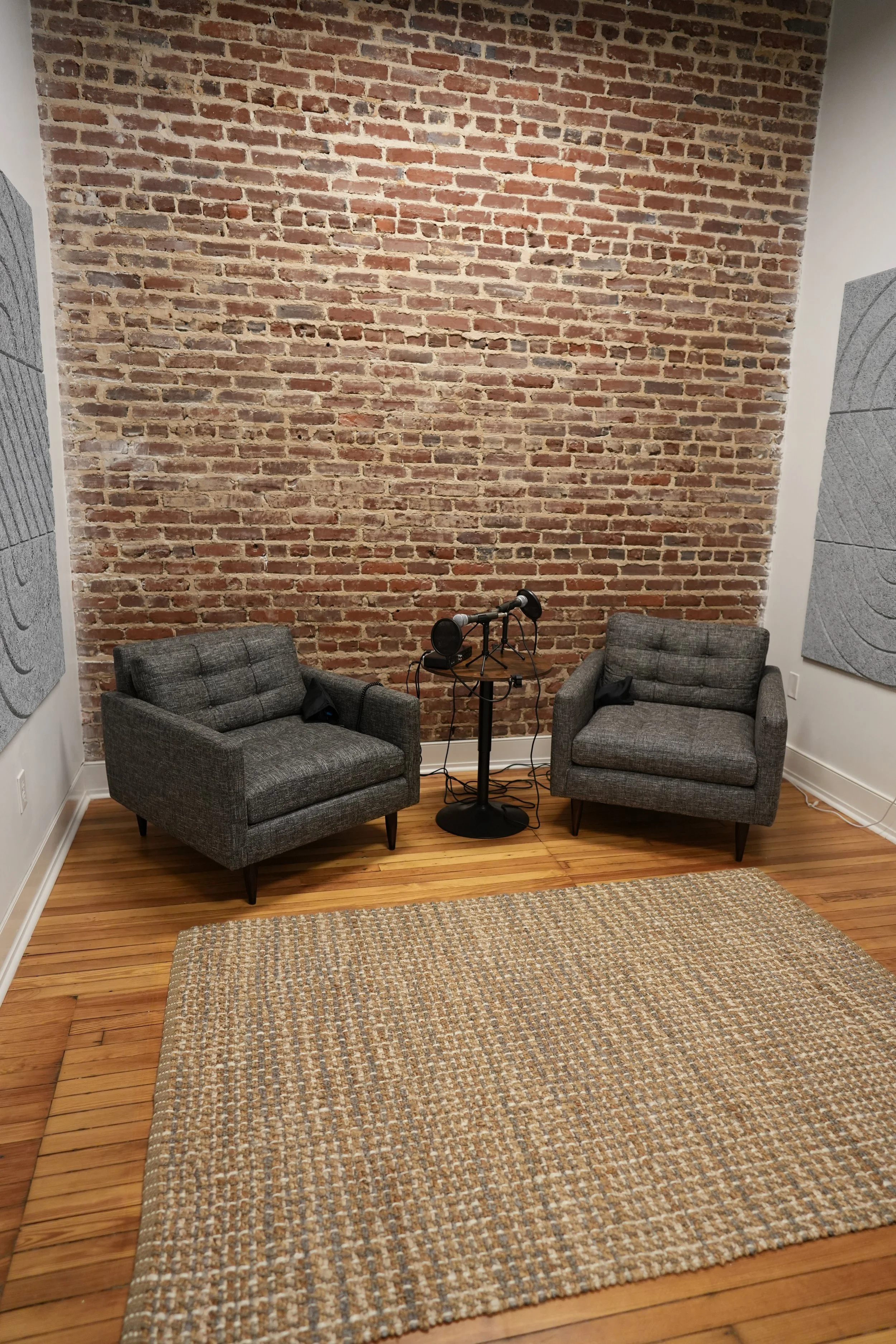 Two gray upholstered armchairs with black wooden legs are positioned in front of an exposed brick wall. Between them is a black microphone on a stand with a pop filter. A beige woven rug covers part of the wooden floor in a cozy, modern room.