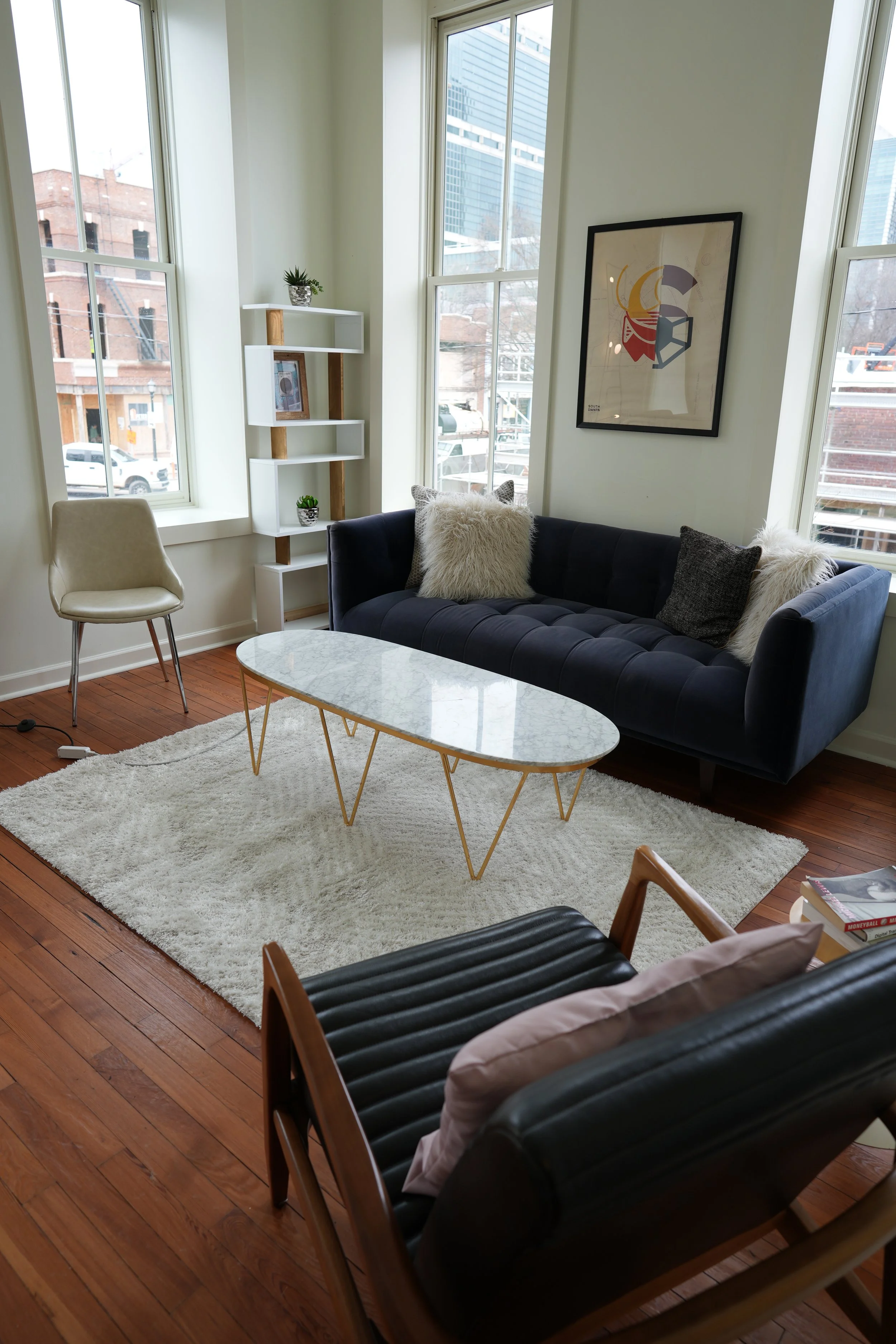 A modern living room with large windows, a navy blue velvet sofa with plush pillows, a white marble coffee table with gold legs, a cream accent chair, a black leather armchair, a white shag rug, and minimalist art and plants.