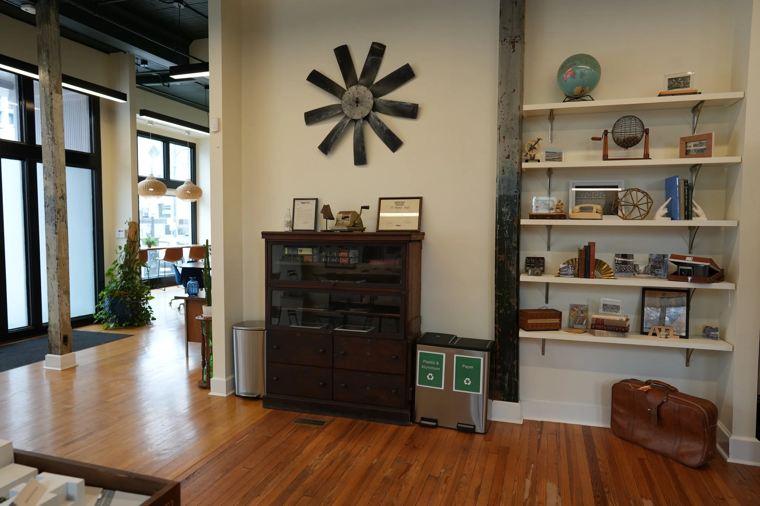 Interior of a modern office or living space with wooden floors, white walls, and large windows. A dark wood cabinet with framed pictures, a vintage telephone, and decorative items sits against the wall. To the right, white shelves hold various decorative objects and framed photos. Two recycling bins labeled "Plastic & Aluminum" and "Paper" are placed beside the cabinet. A brown leather bag rests on the floor nearby. Part of a dining or conference area with chairs and a table can be seen in the background, along with some plants and pendant lighting.