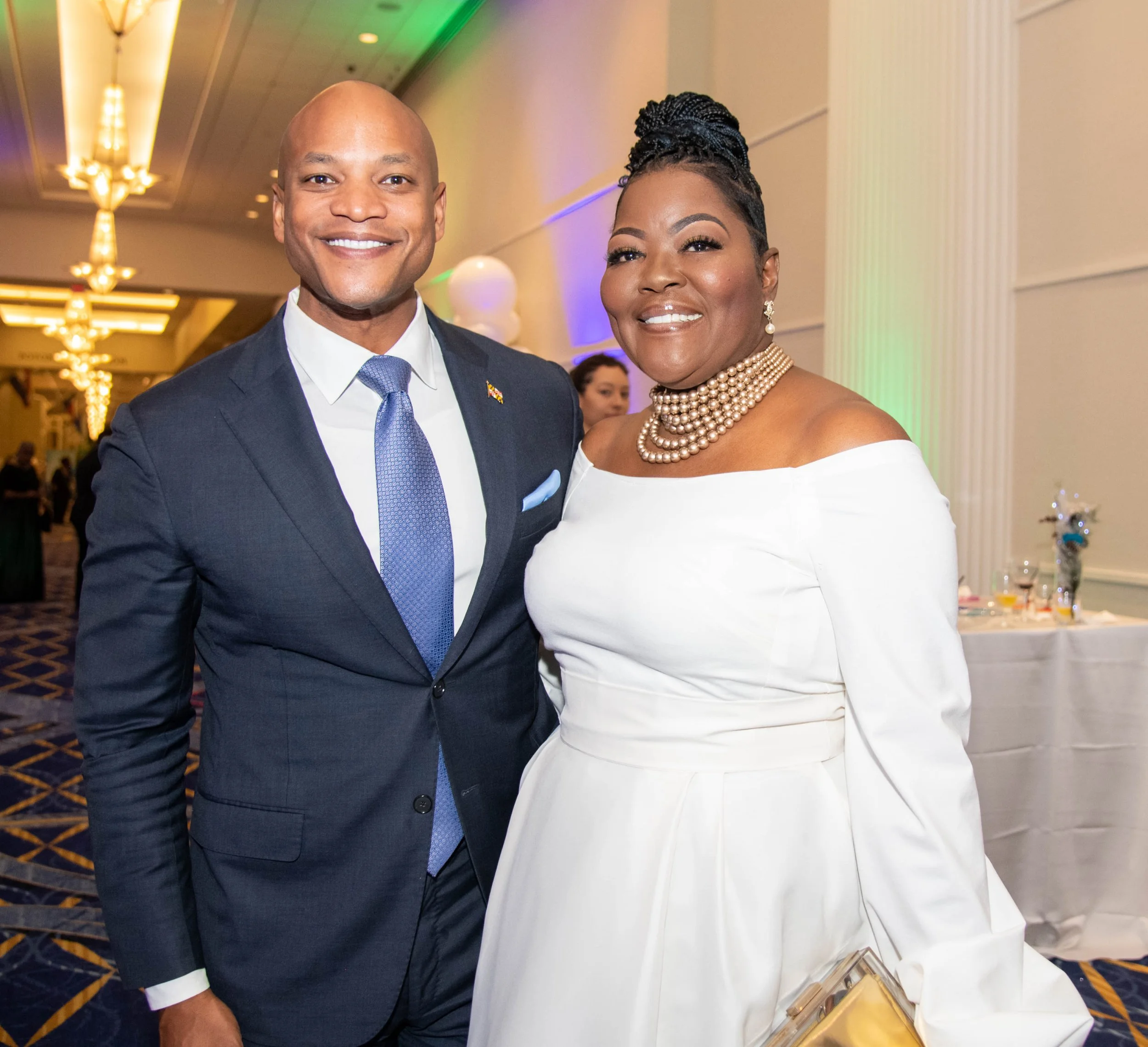 A smiling man in a navy suit with a blue tie standing next to a smiling woman in a white off-shoulder dress with pearl jewelry at an event.