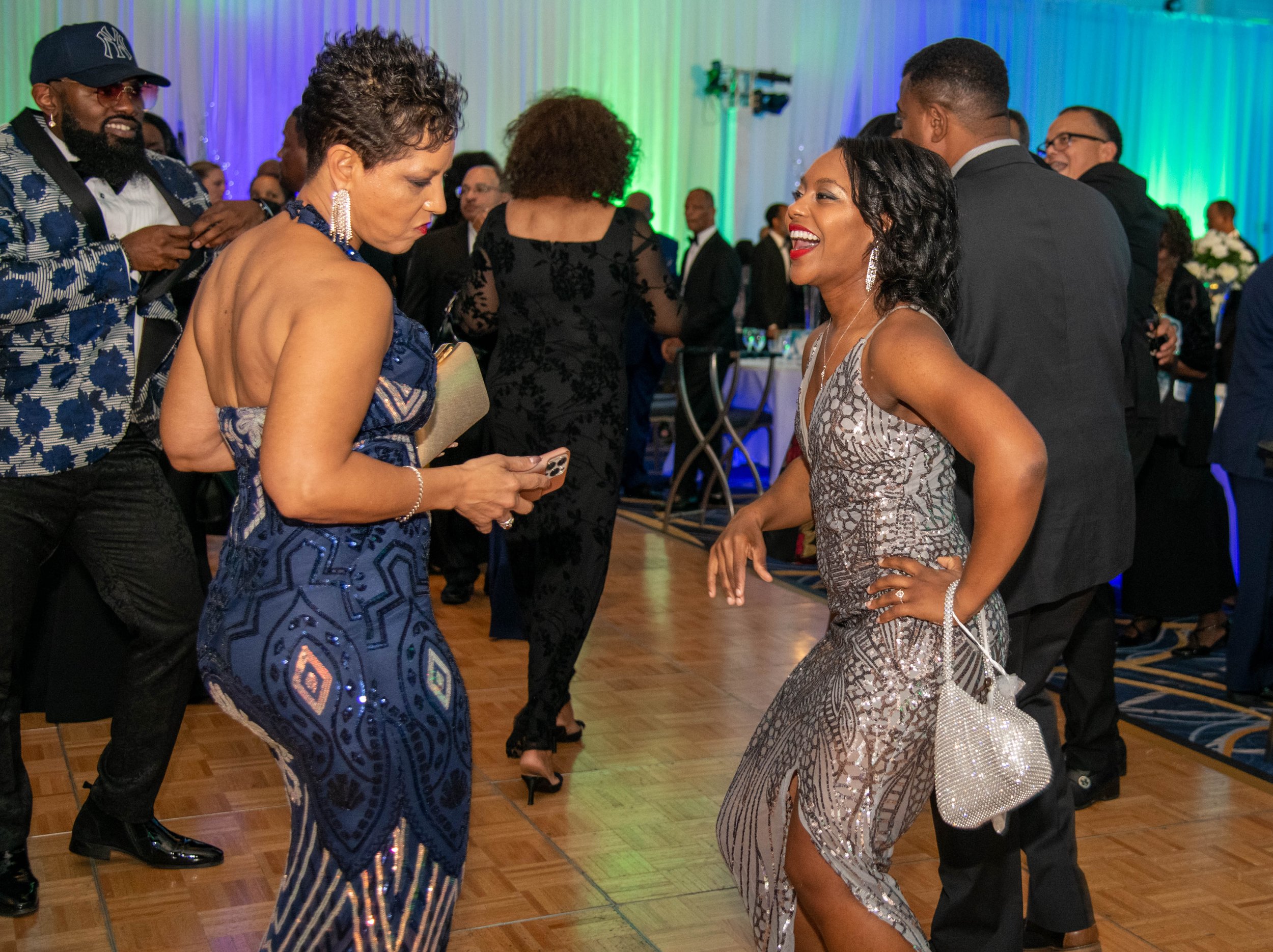 Two women dancing and talking at a formal event, surrounded by other guests in elegant attire, with colorful lighting and a decorated background.