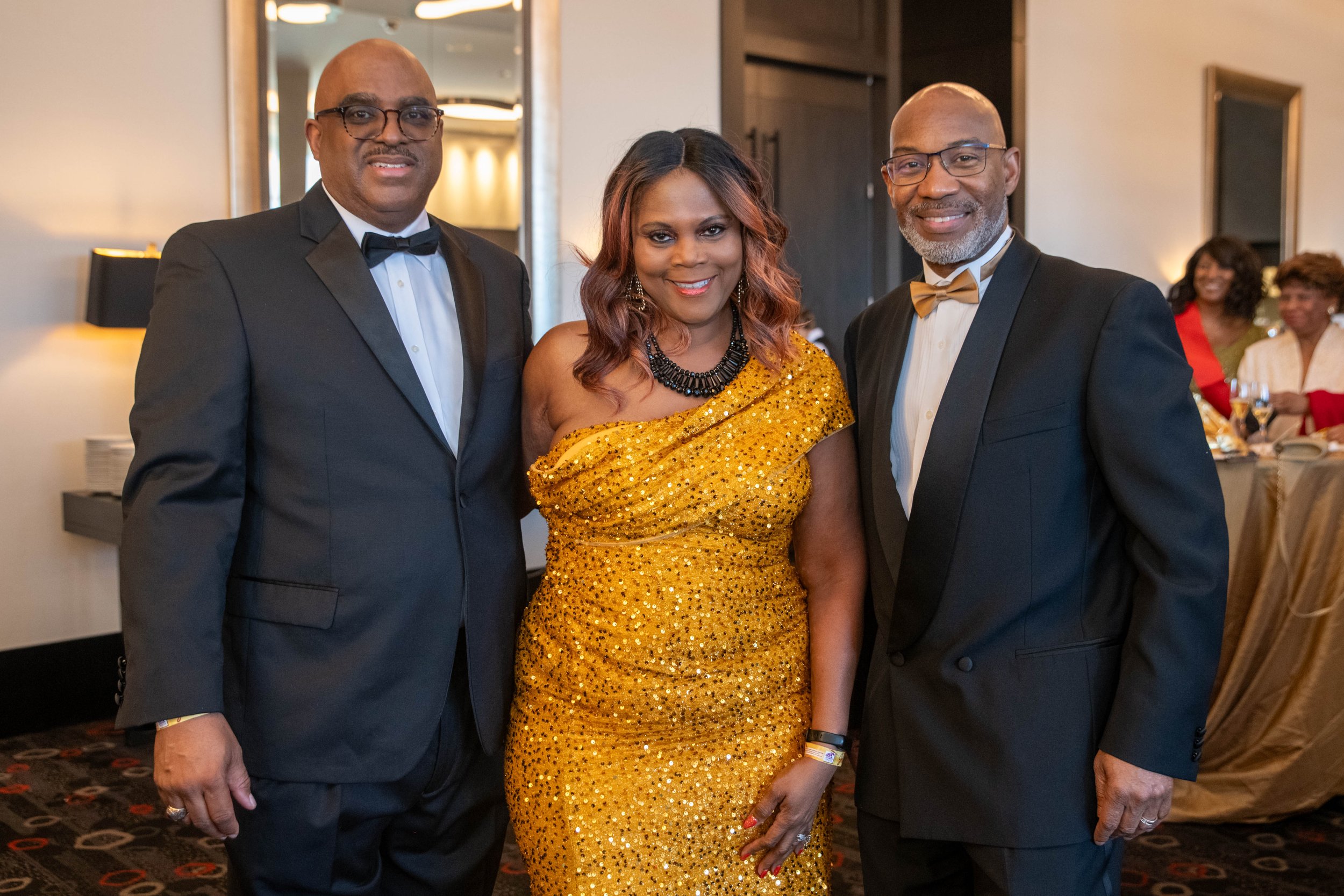 Three well-dressed individuals at an event, two men in tuxedos with glasses and a woman in a gold sequin gown, standing closely together and smiling.