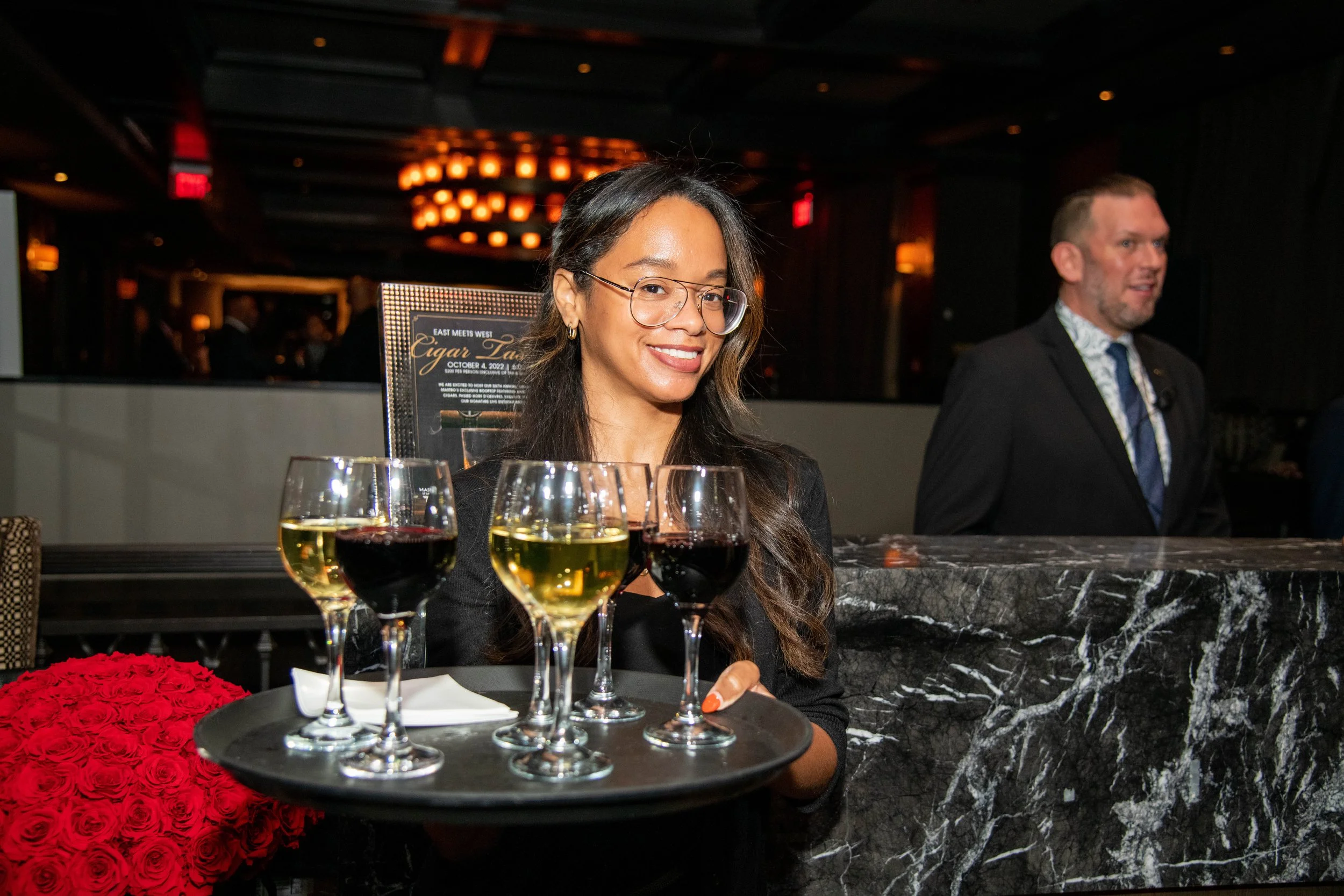A woman with glasses, long dark hair, and wearing a black blazer, smiling, is holding a tray with five glasses of white and red wine. Behind her, a man in a dark suit and patterned tie stands at a marble bar in a dimly lit restaurant or bar. There is