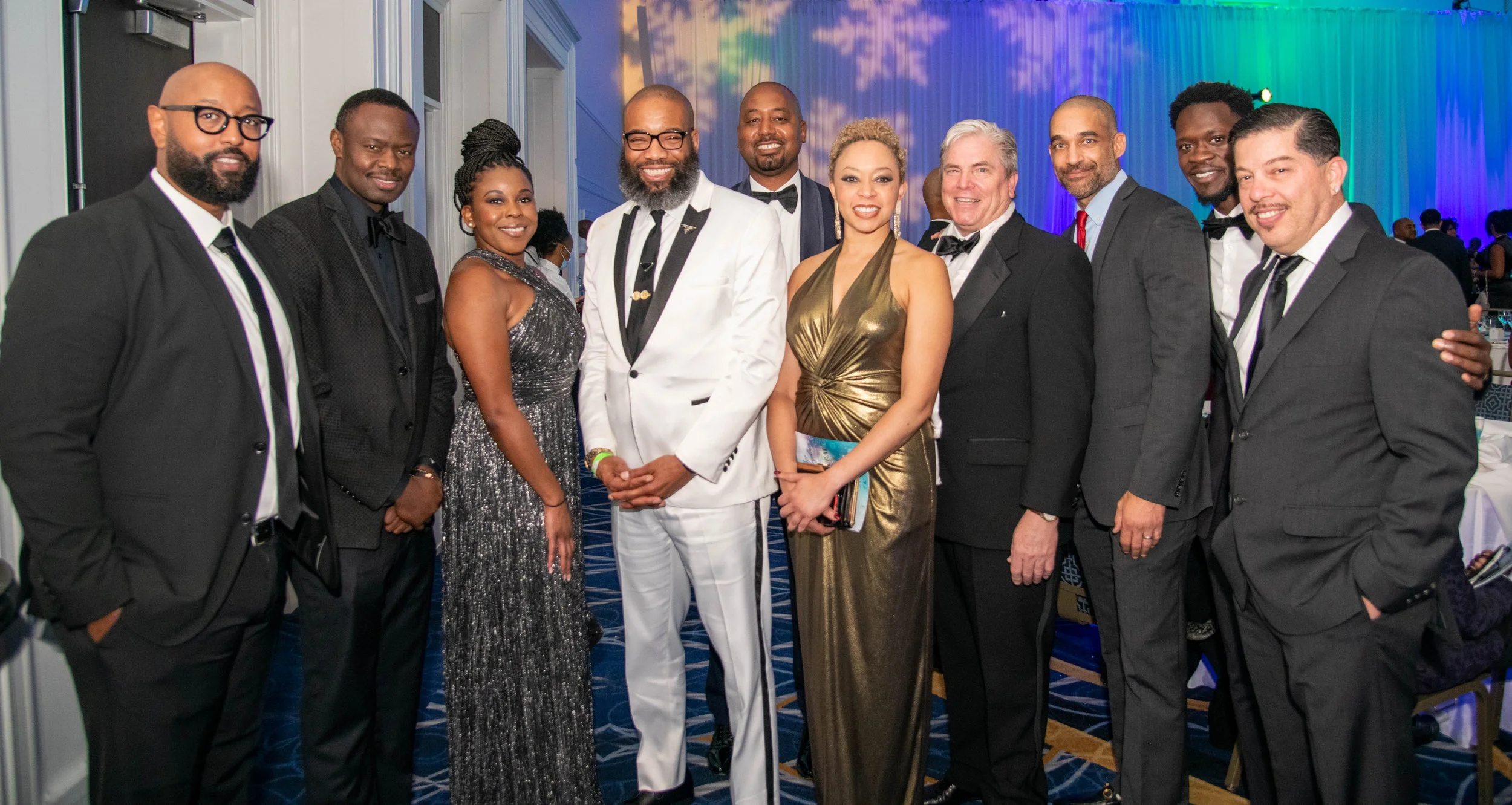 Group of twelve people dressed in formal attire standing together at an event with colorful lighting in the background.
