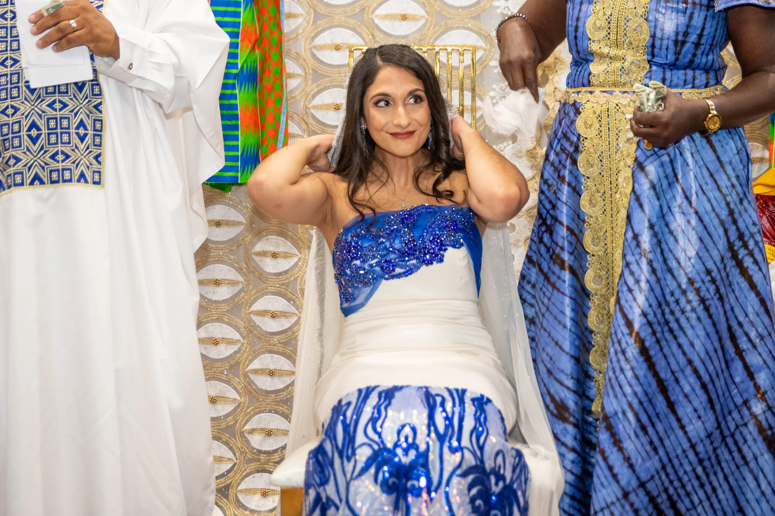 Bride in a white and blue dress sitting on a chair surrounded by men in colorful traditional African attire during a traditional African wedding.