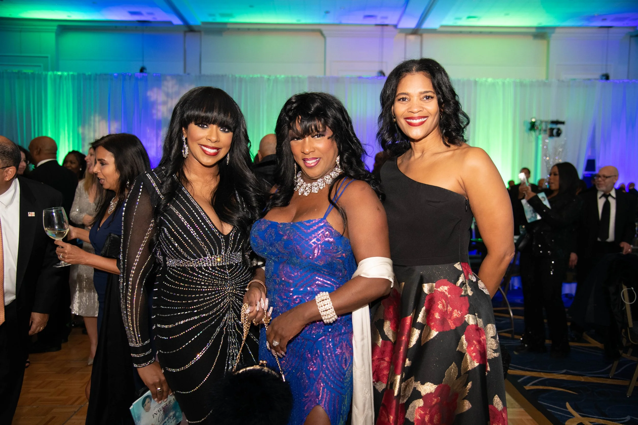 Three women dressed in evening gowns at a formal event, smiling for the camera. The background has colorful lights and other guests socializing.